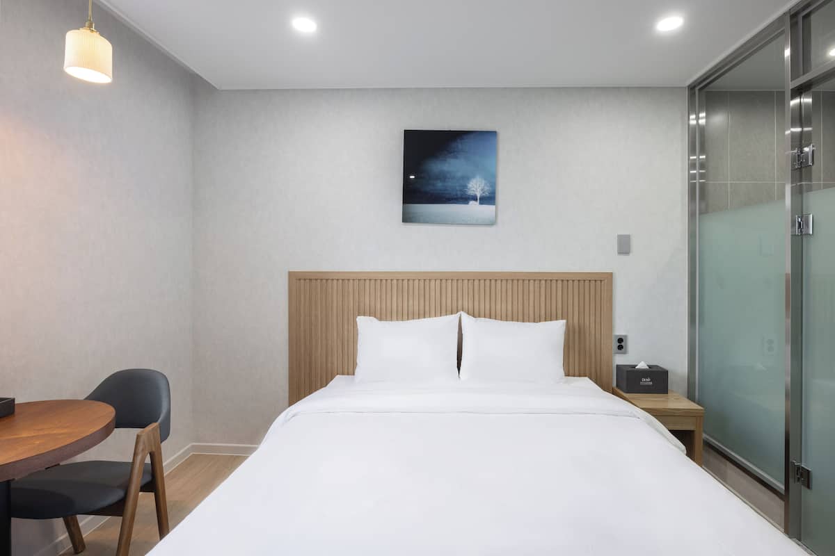 A hotel room with a bed, a wooden headboard, a nightstand, a desk with a chair, and a painting on the wall.