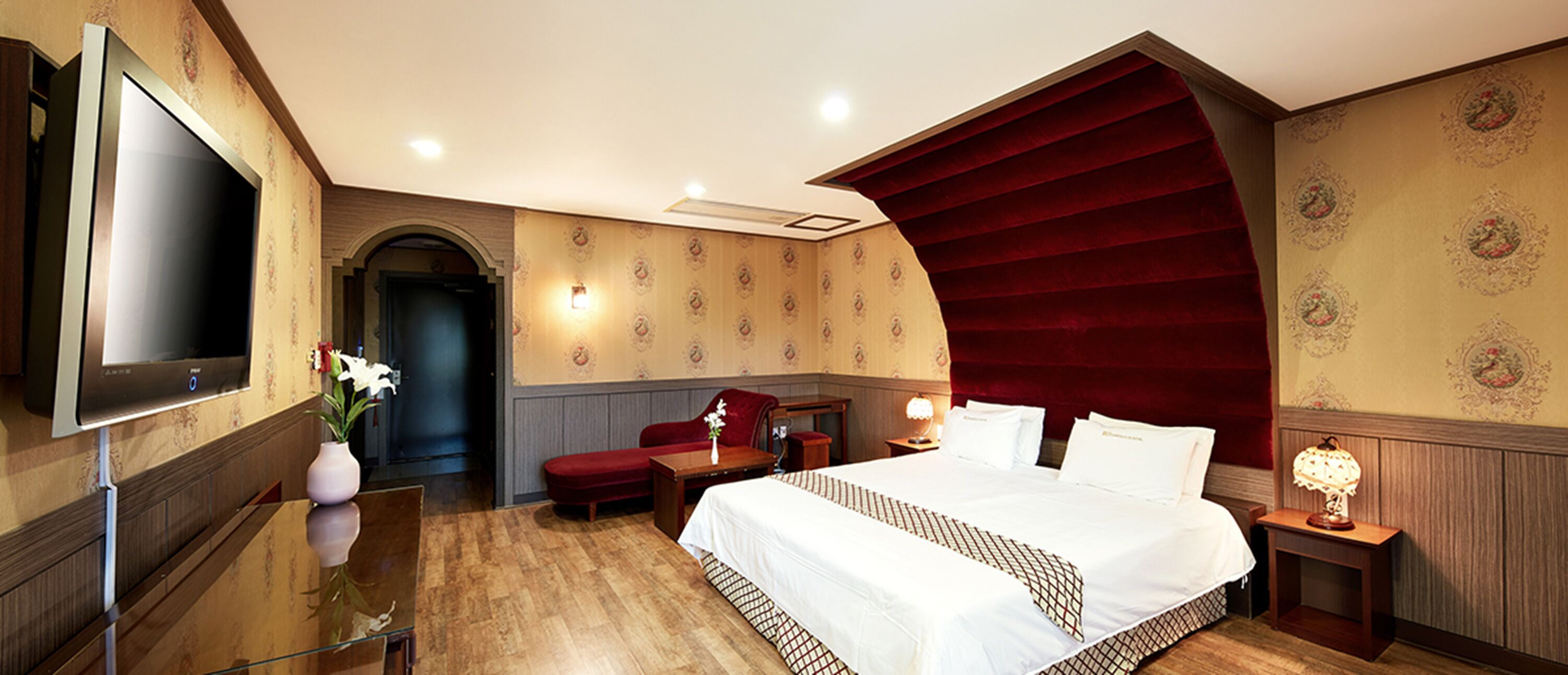 A hotel room with a large bed, a desk, a chair, a TV, and a red velvet wall.