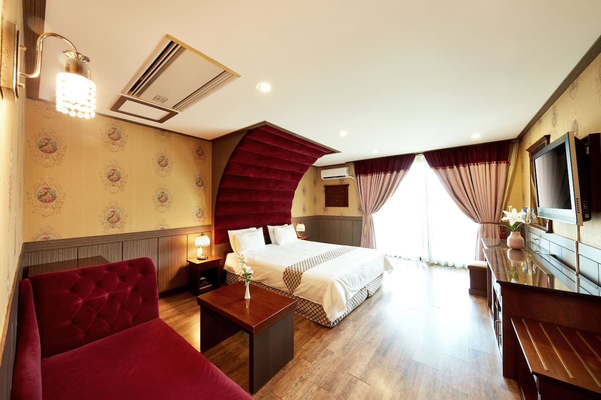 A hotel room with a bed, a red sofa, a wooden desk, a TV, and a large window with curtains.