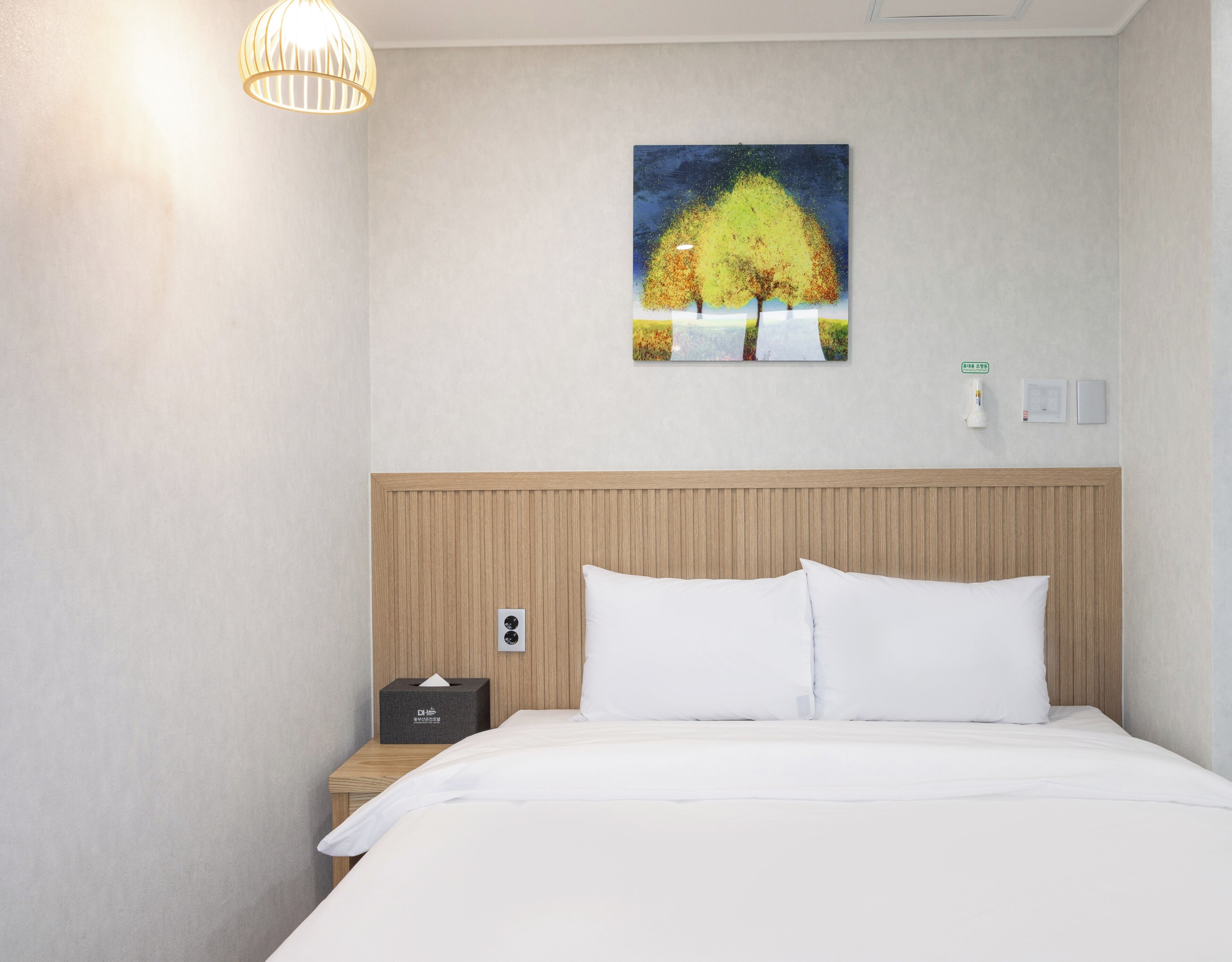 A hotel room with a wooden headboard, a white bed, a nightstand with a tissue box, a wall-mounted painting of trees, and a ceiling light fixture.