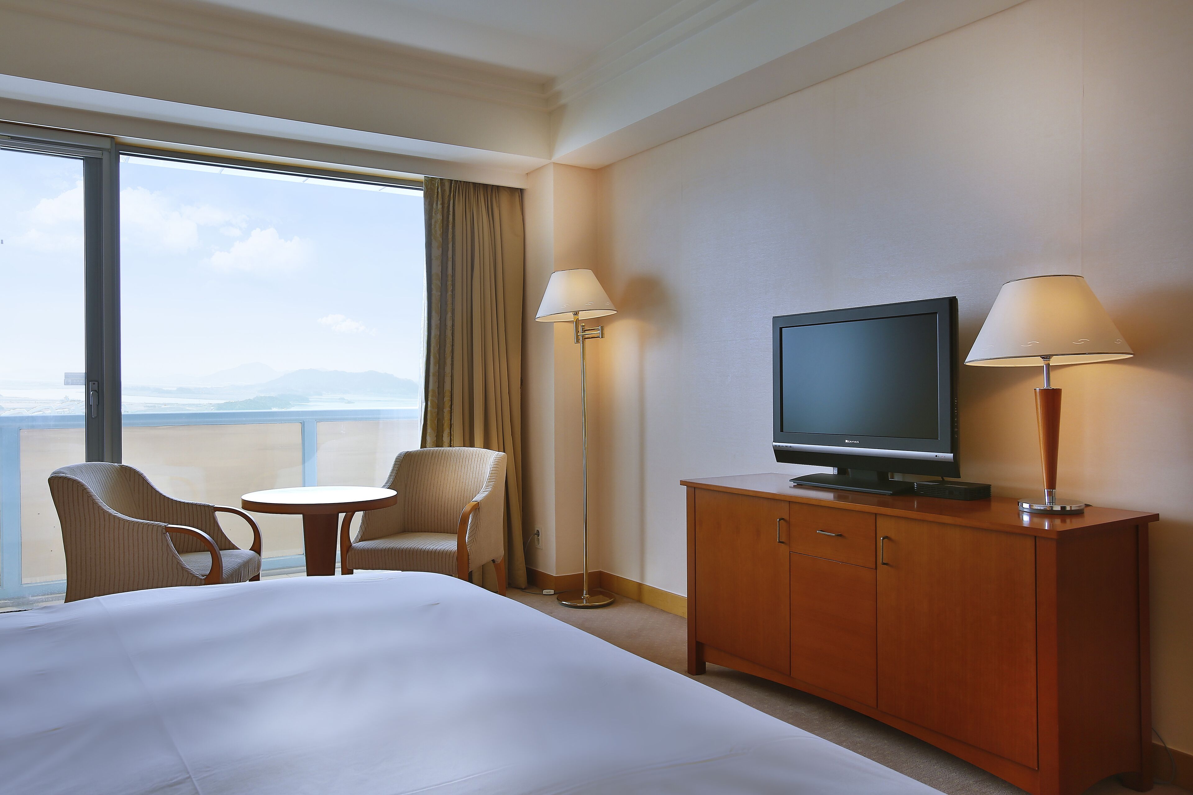 Quarto de hotel com uma cama, duas cadeiras, uma mesinha, uma televisão sobre um armário de madeira e uma janela ampla com vista para o mar.