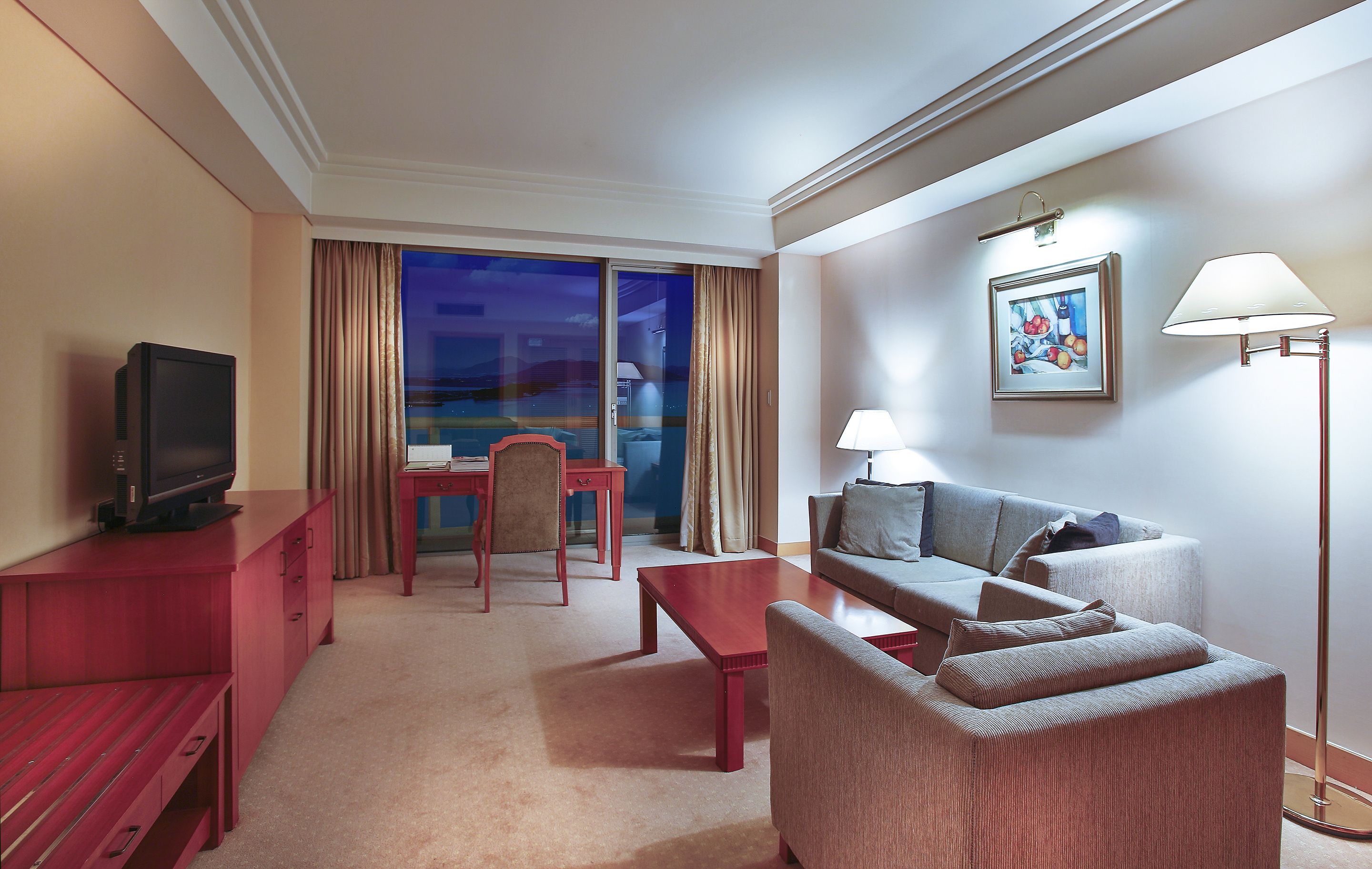 A hotel room with a television, a sofa, a chair, a table, and a floor lamp.