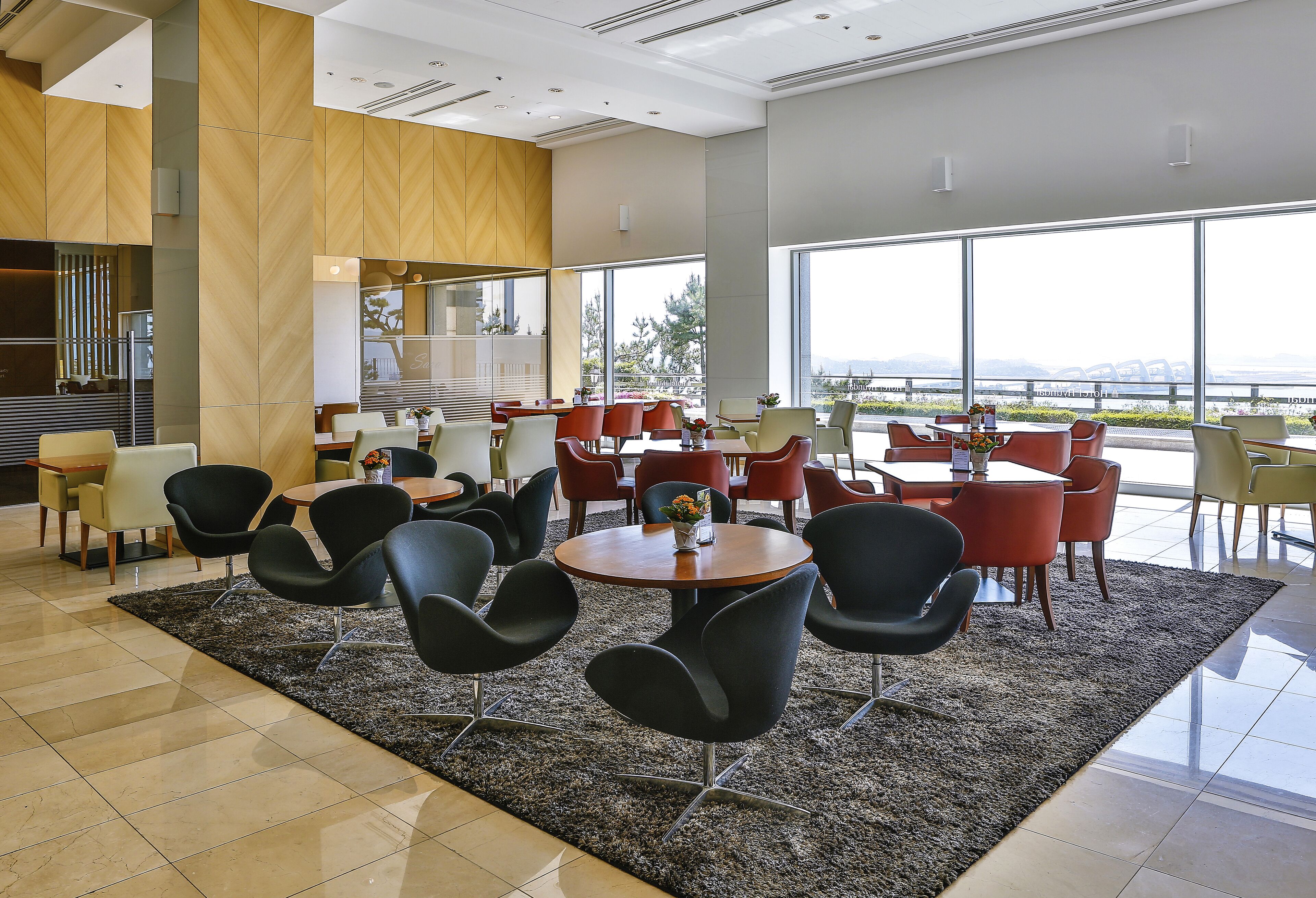 A modern dining area with round tables and black chairs, large windows with a view, and a mix of red and yellow chairs.