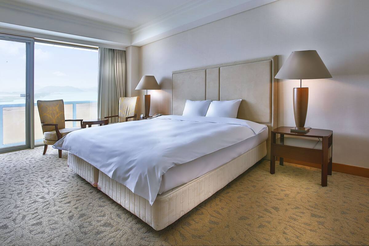 Ein Hotelzimmer mit einem großen Bett, zwei Nachttischen mit Lampen, einem Sessel und Blick auf den Ozean durch ein großes Fenster.