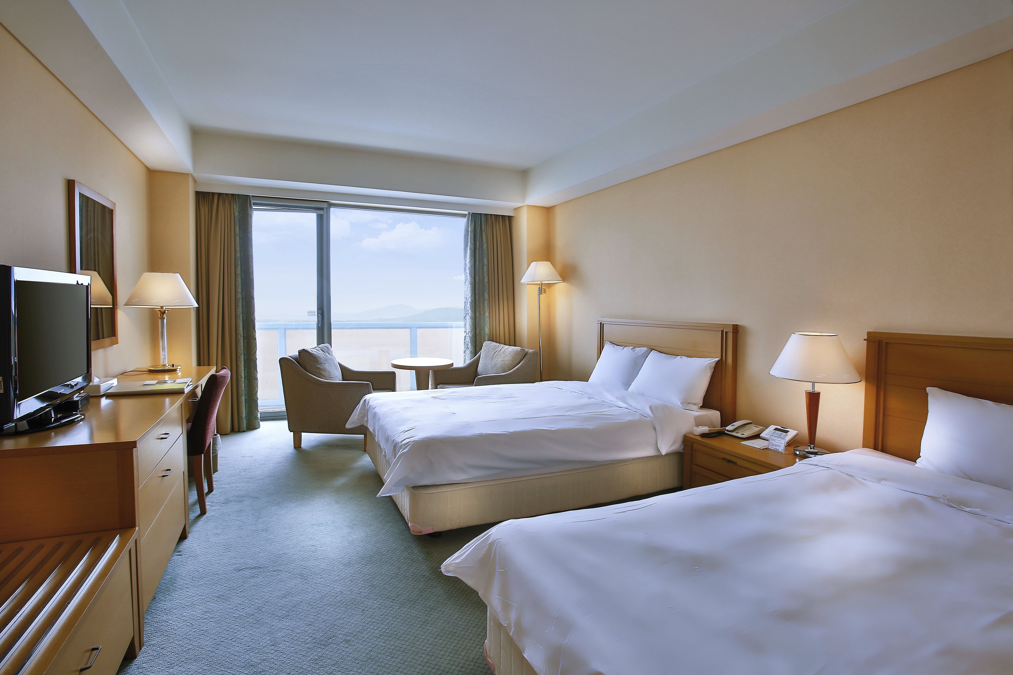 Ein Hotelzimmer mit zwei Betten, einem Schreibtisch mit Fernseher, einem Sessel, einem kleinen Tisch und Meerblick.