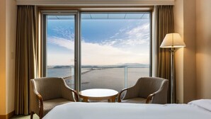 Ein Hotelzimmer mit großem Fenster, zwei Sesseln, einem kleinen runden Tisch und einem Bett.