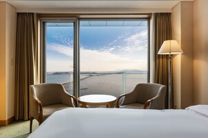 Una habitación de hotel con un ventanal grande, dos sillones, una mesita redonda y una cama.