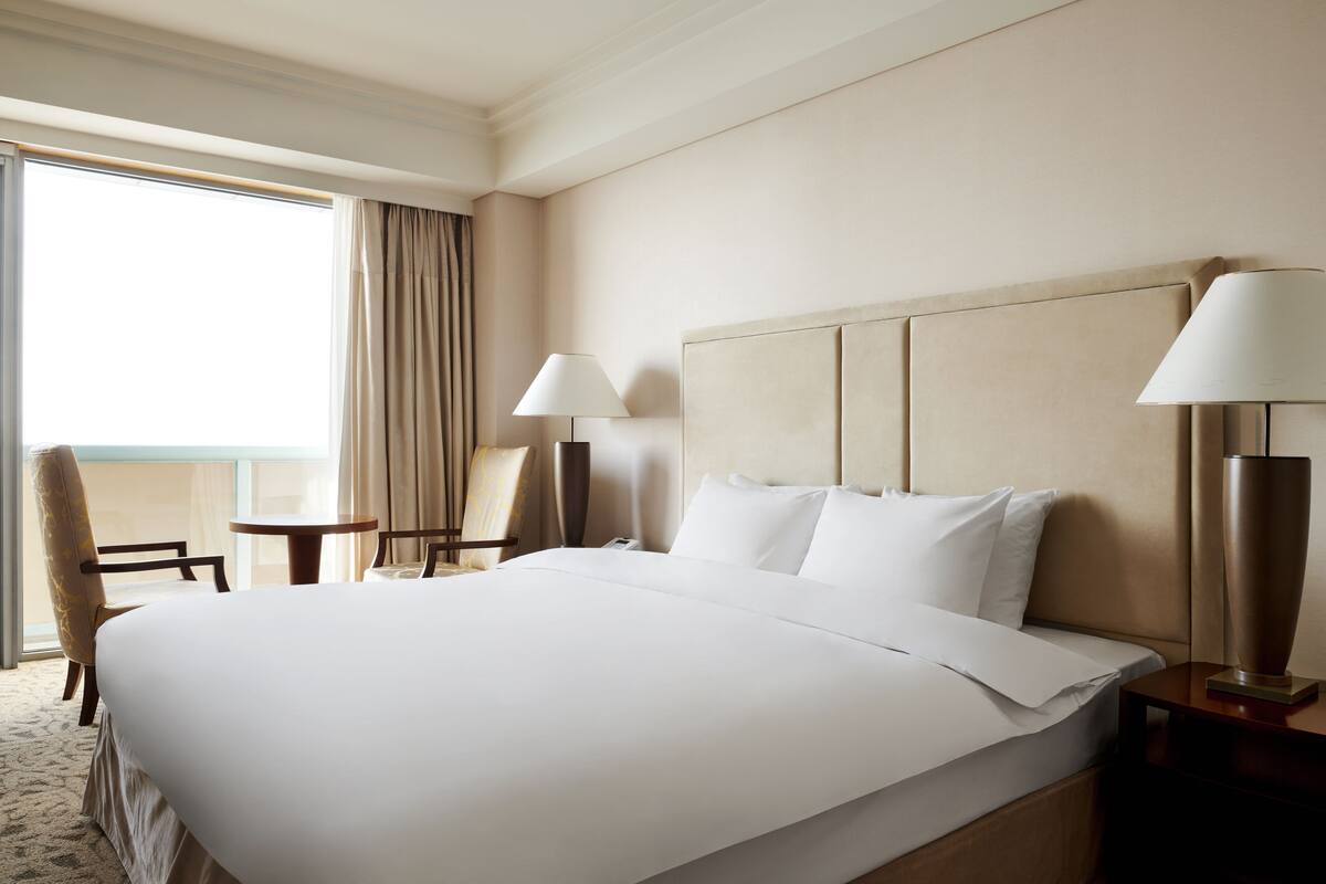 Ein Hotelzimmer mit einem großen Bett, zwei Nachttischen, einem Sessel und Blick ins Freie.