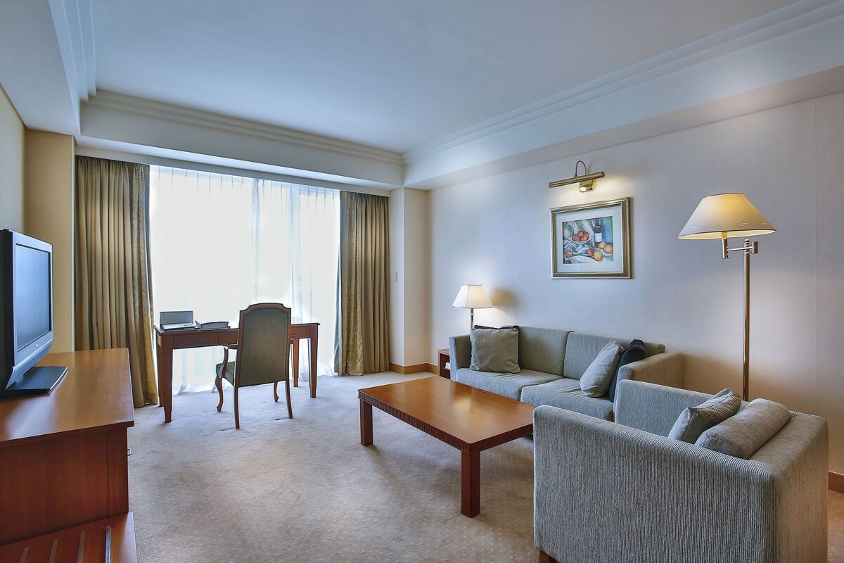 Ein Hotelzimmer mit Flachbildfernseher, Schreibtisch aus Holz, einem Laptop, einer grauen Couch, einem Couchtisch und einer Lampe.