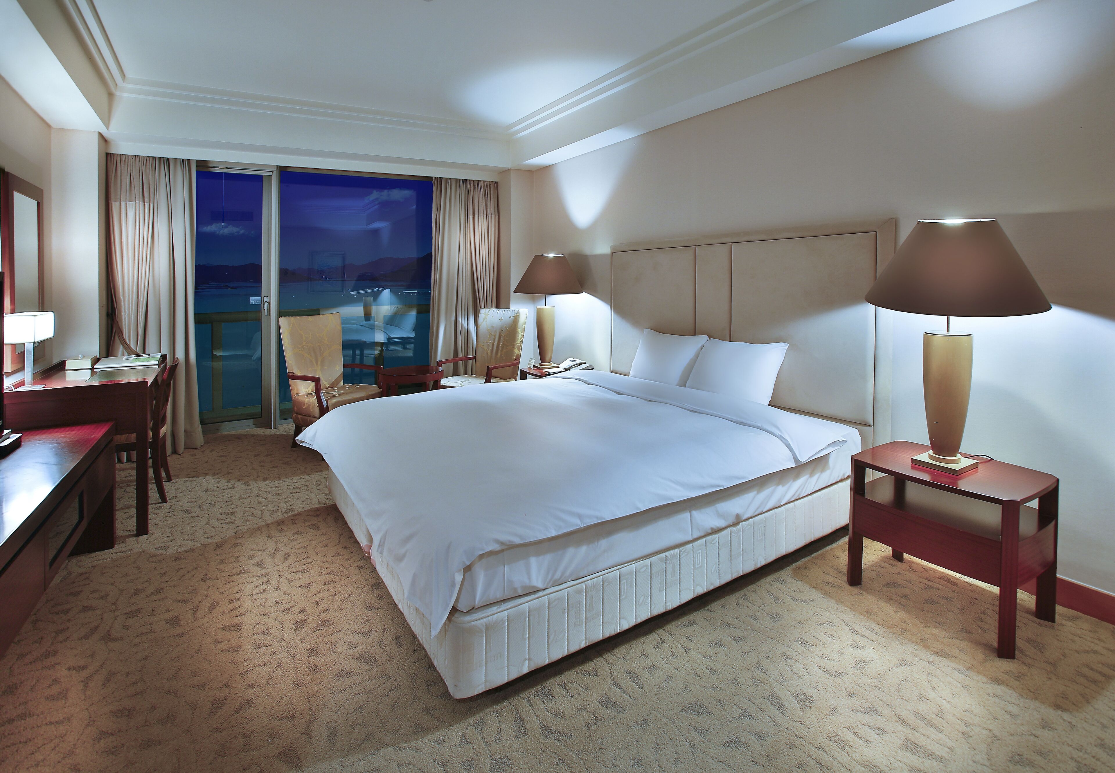 A hotel room with a large bed, two bedside tables with lamps, a desk, and a chair.