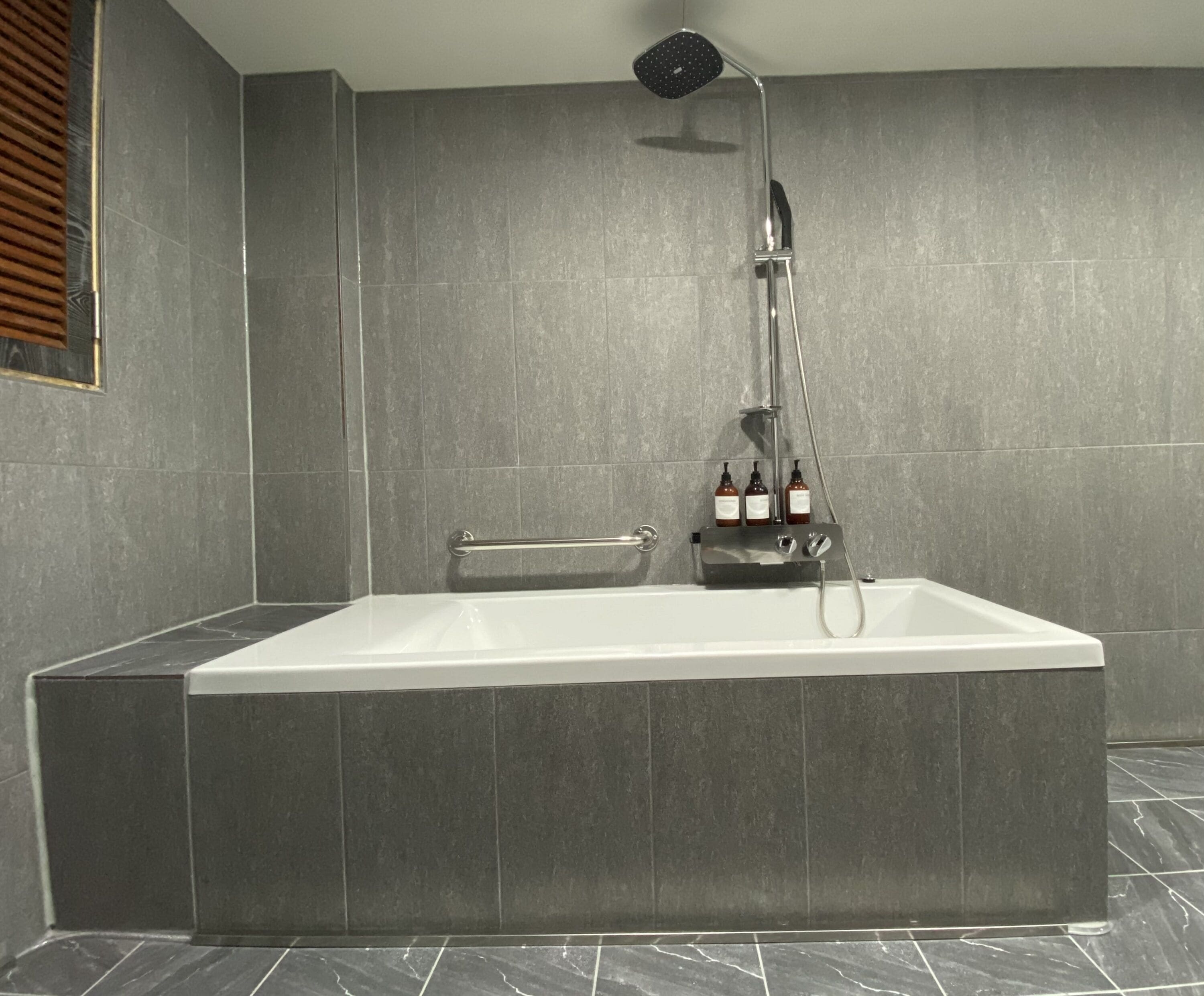 A modern bathroom with a large rectangular bathtub, a handheld showerhead, and two bottles of liquid soap on a shelf.