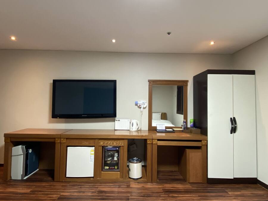A hotel room with a large flat-screen TV, a minibar, a refrigerator, a trash can, a mirror, and a wardrobe.