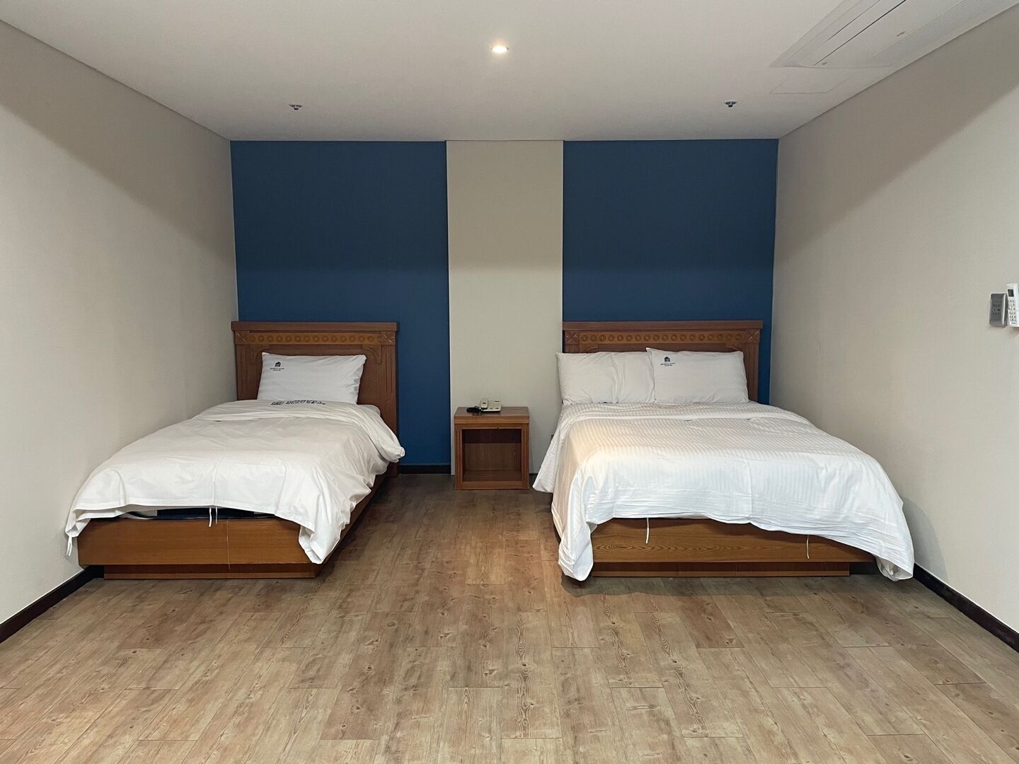 A hotel room with two single beds, wooden headboards, a small bedside table with a lamp, and a plain white wall.