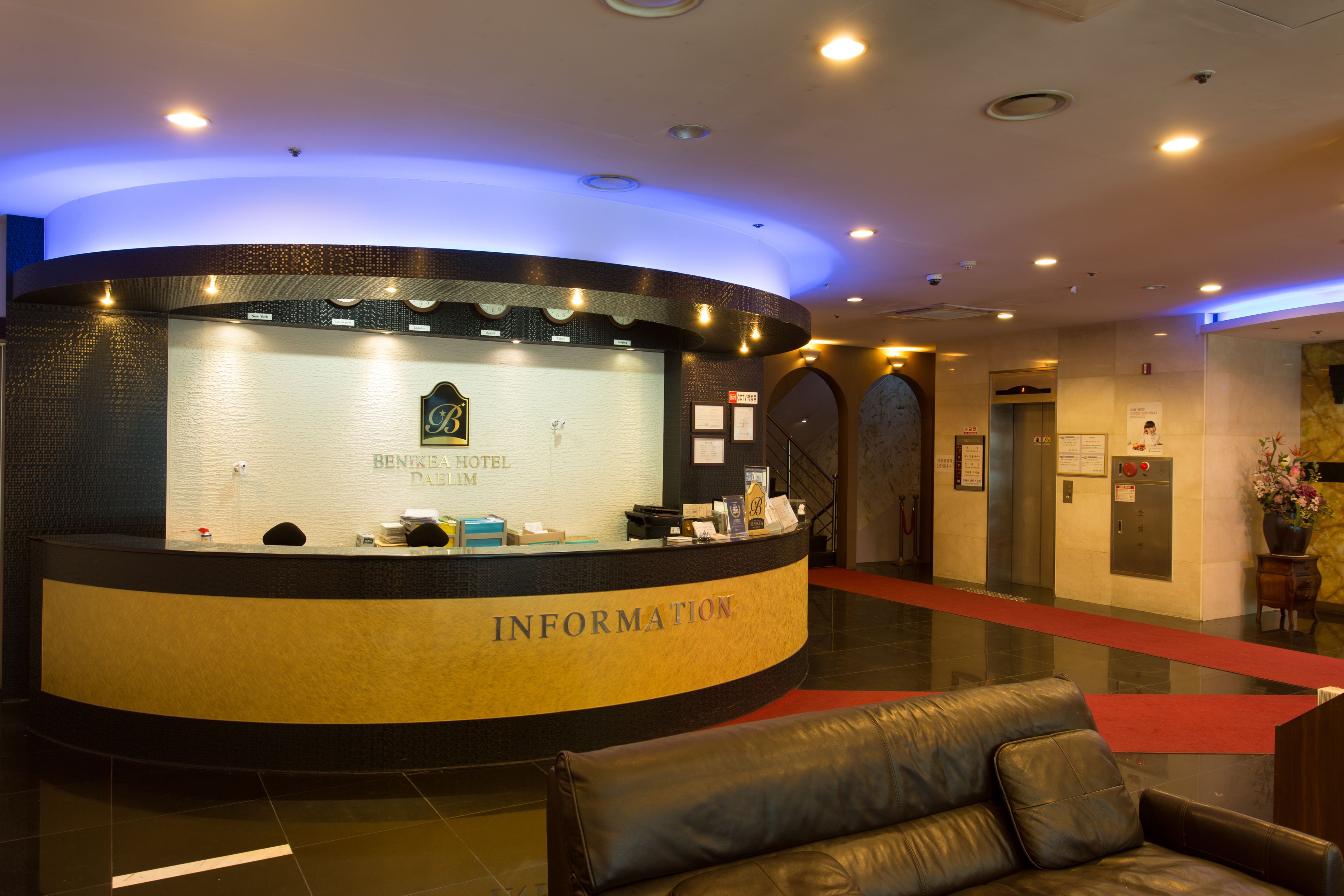 A hotel reception area with a curved reception desk labeled "INFORMATION," a reception desk with a sign for "BENKEA HOTEL DAIHIM," and a red carpet leading to an elevator.