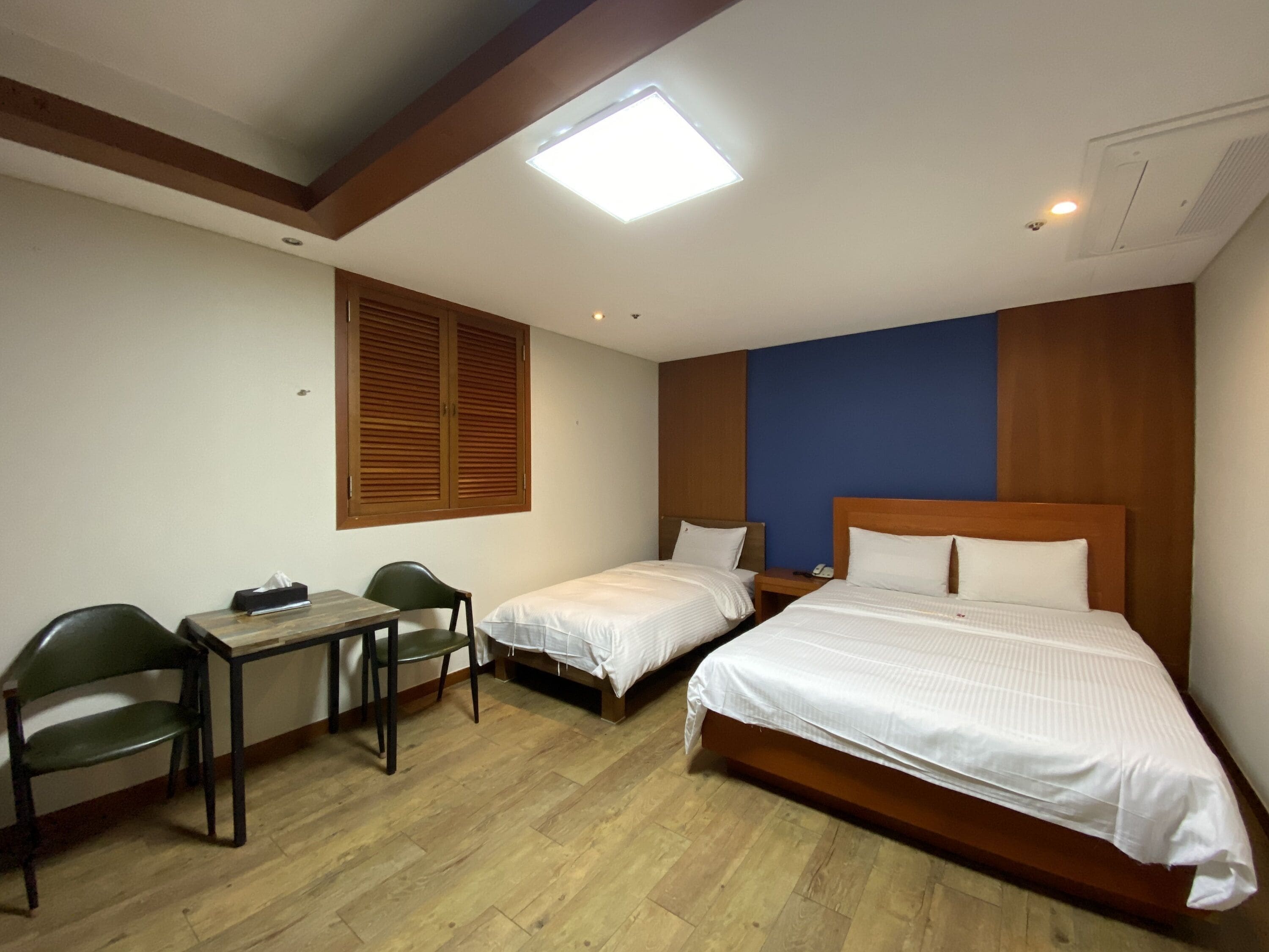 A hotel room with two single beds, a desk with a chair, a wooden floor, and a large window with blinds.