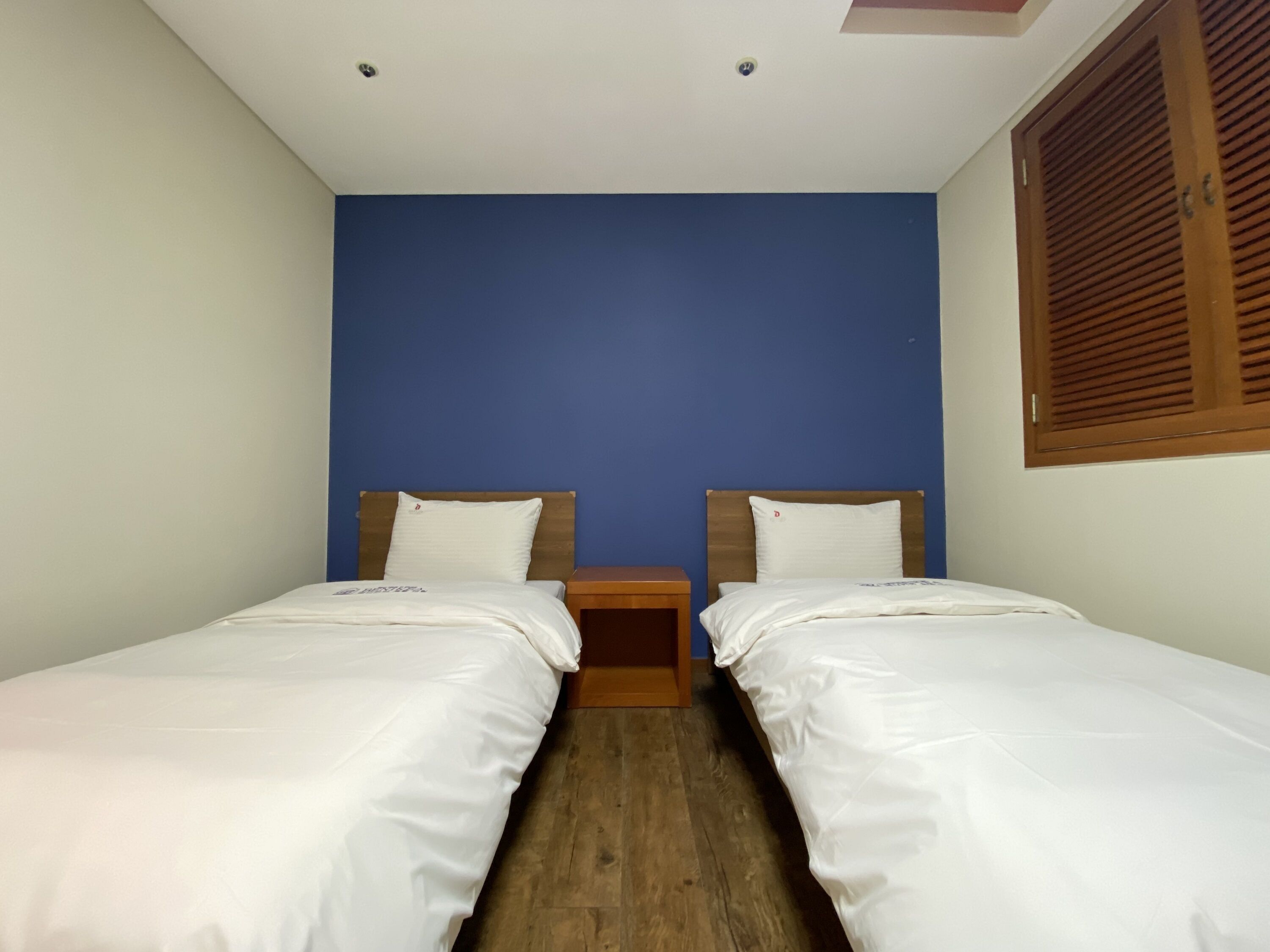 A room with two single beds, wooden headboards, a nightstand between them, and a window with wooden shutters.