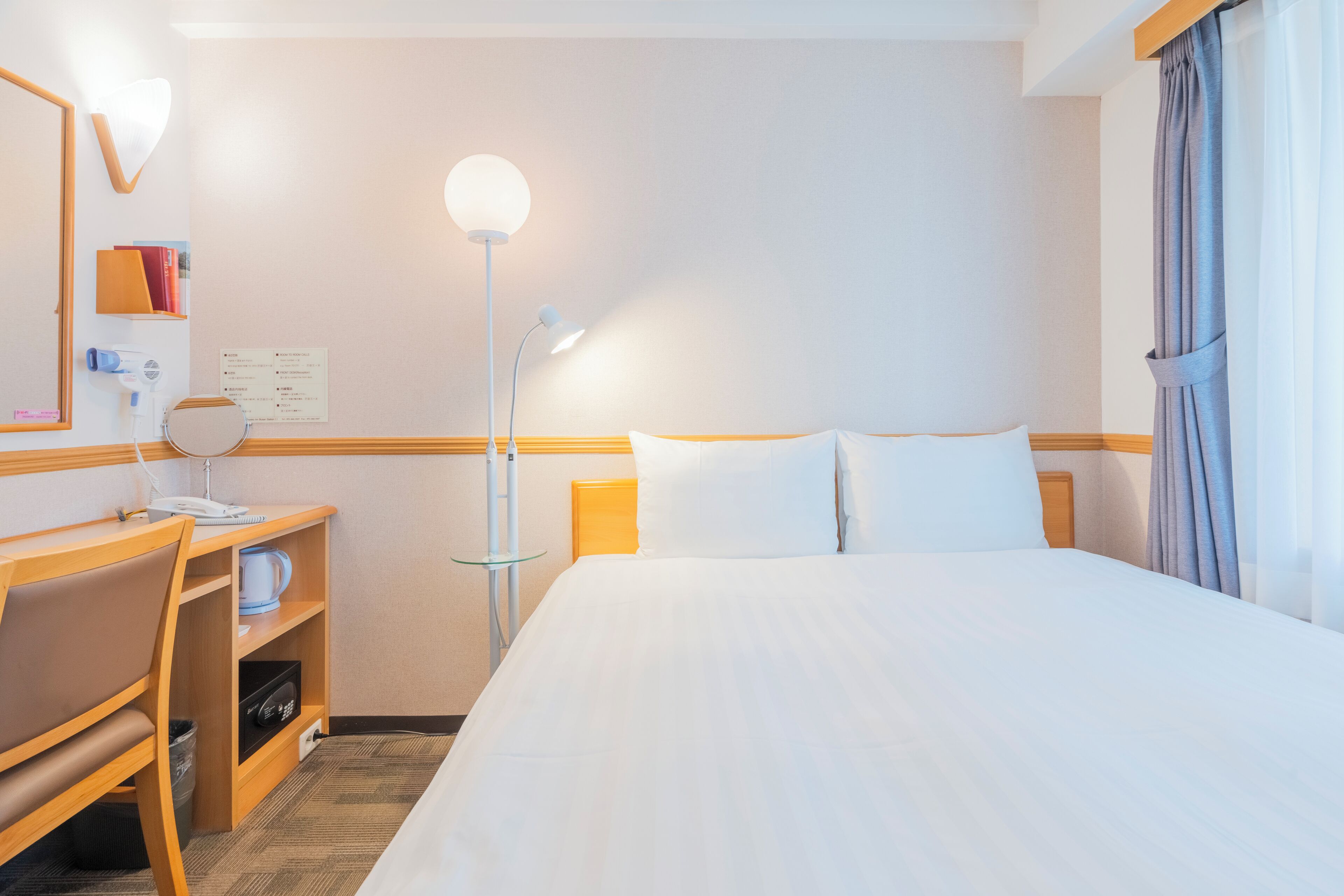 A hotel room with a bed, a desk with a chair, a lamp, and a mirror.
