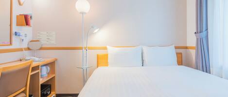 A hotel room with a bed, a desk with a chair, a lamp, and a mirror.