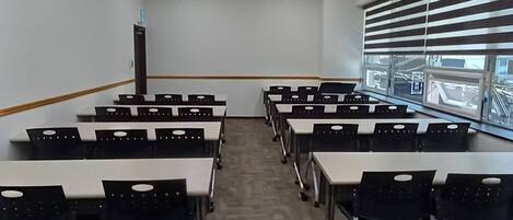 Une salle de classe avec des rangées de bureaux, un projecteur, une horloge et une fenêtre avec des stores.