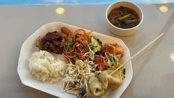 A plate of food including rice, noodles, vegetables, and dumplings, with a bowl of soup on a table.