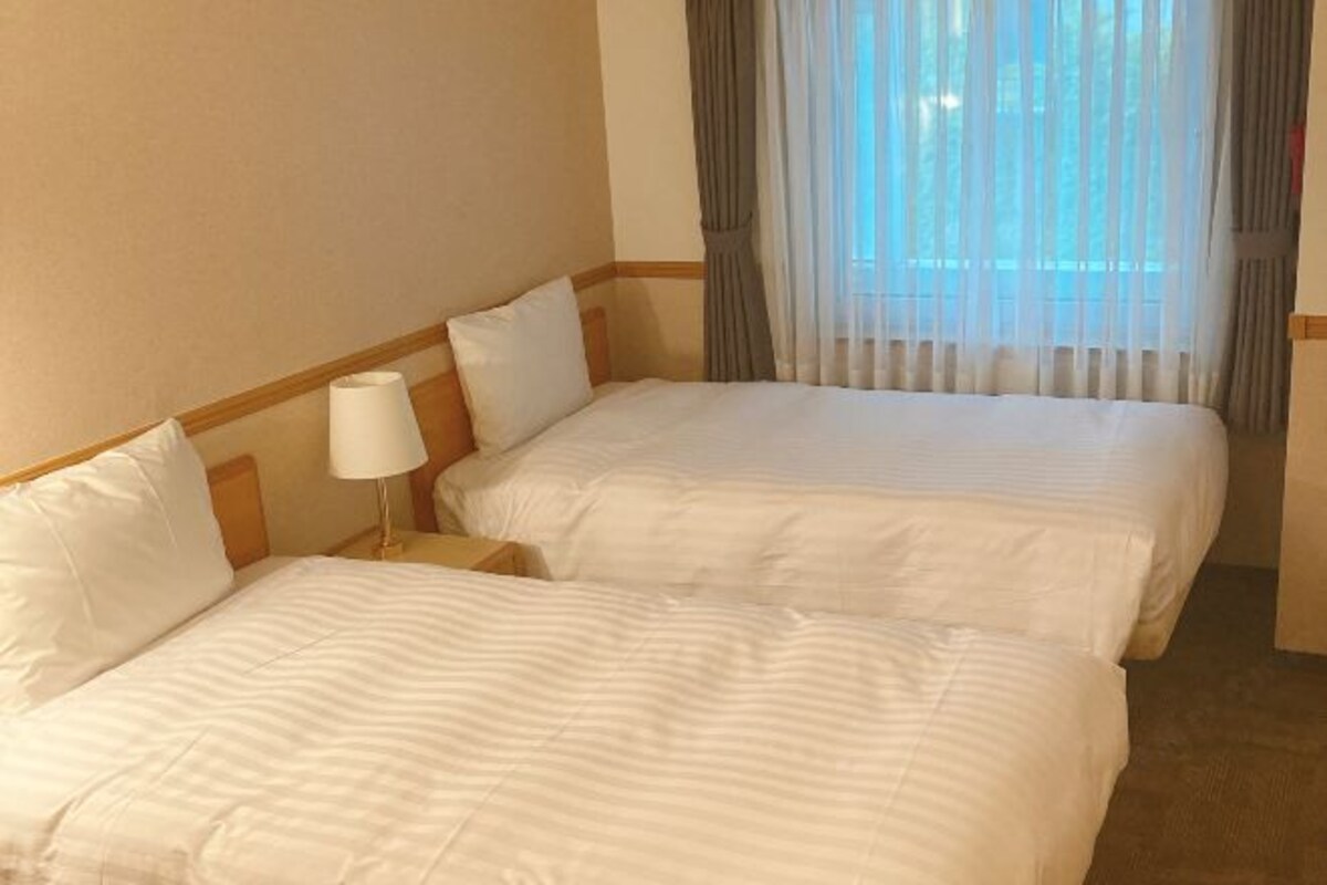 A hotel room with two beds, a nightstand, a lamp, and sheer curtains.