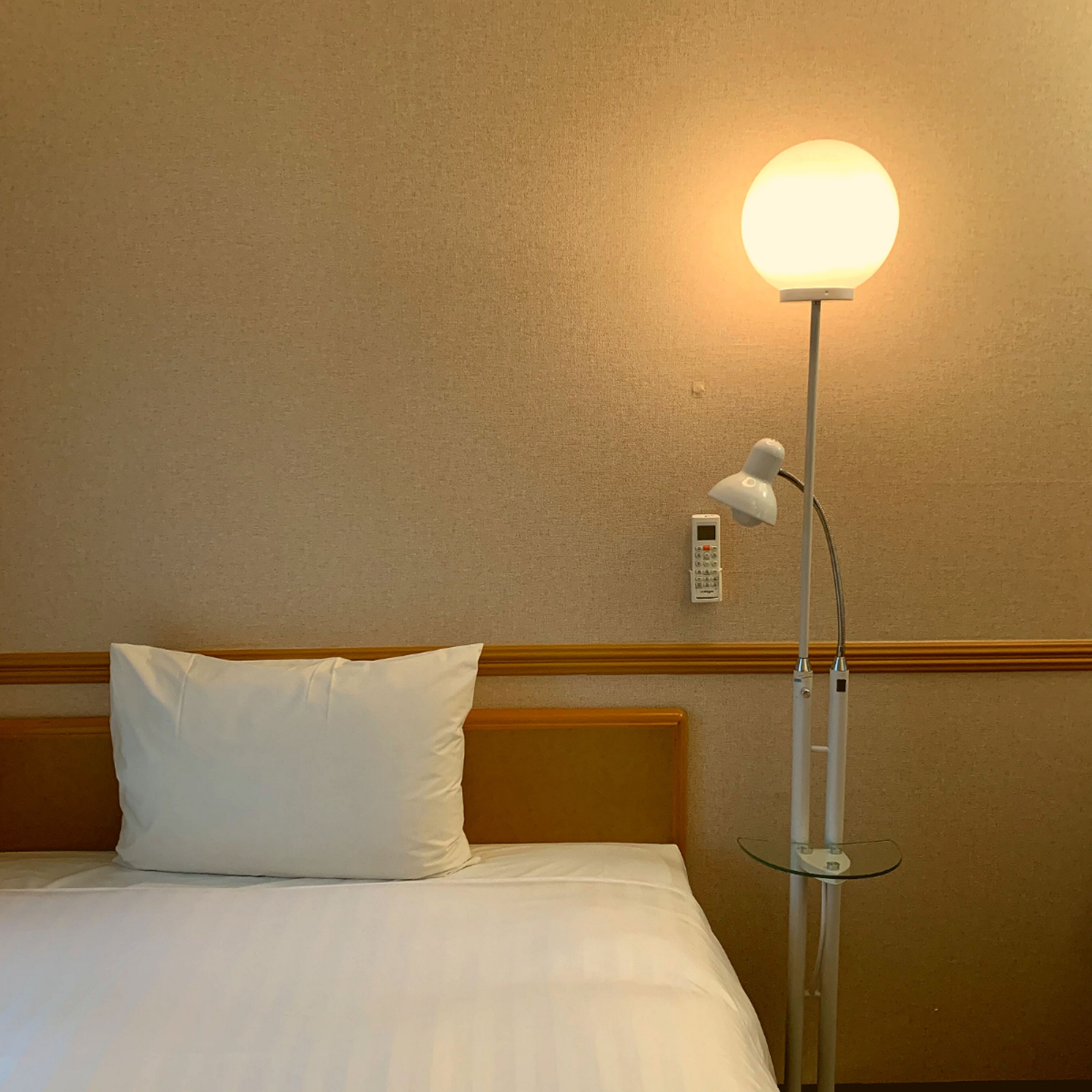 Una habitación de hotel con una cama, un almohadón blanco, una mesita de noche, una lámpara y un teléfono en la pared.