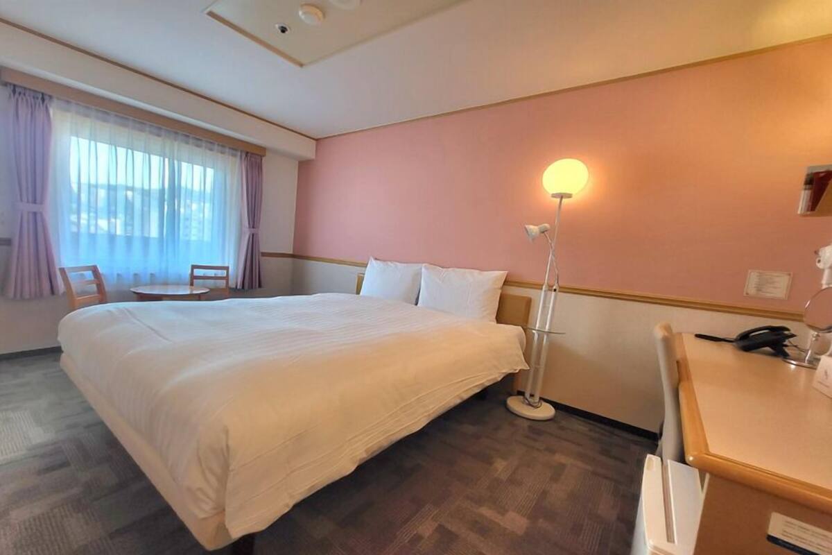 A hotel room with a large bed, a desk, a chair, a desk lamp, and a window with curtains.