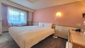 A hotel room with a large bed, a desk, a chair, a desk lamp, and a window with curtains.