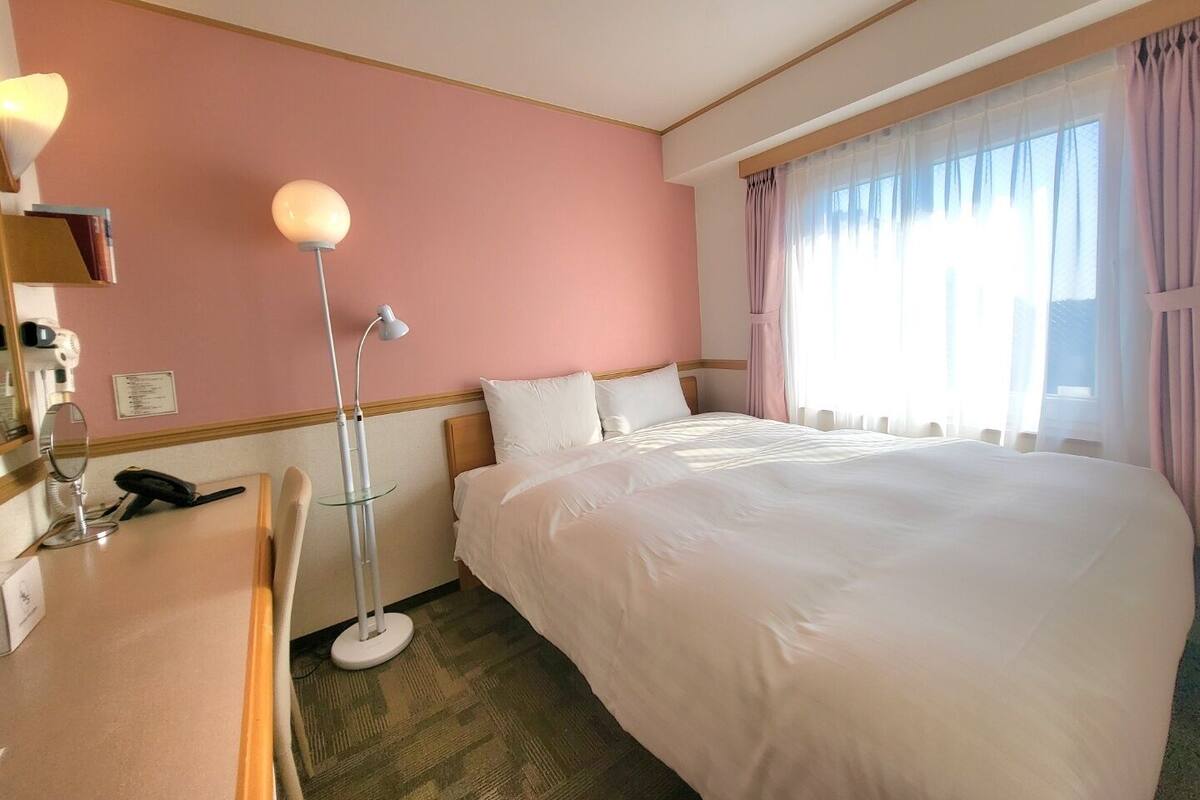 A hotel room with a large bed, a desk with a lamp, and a window with curtains.