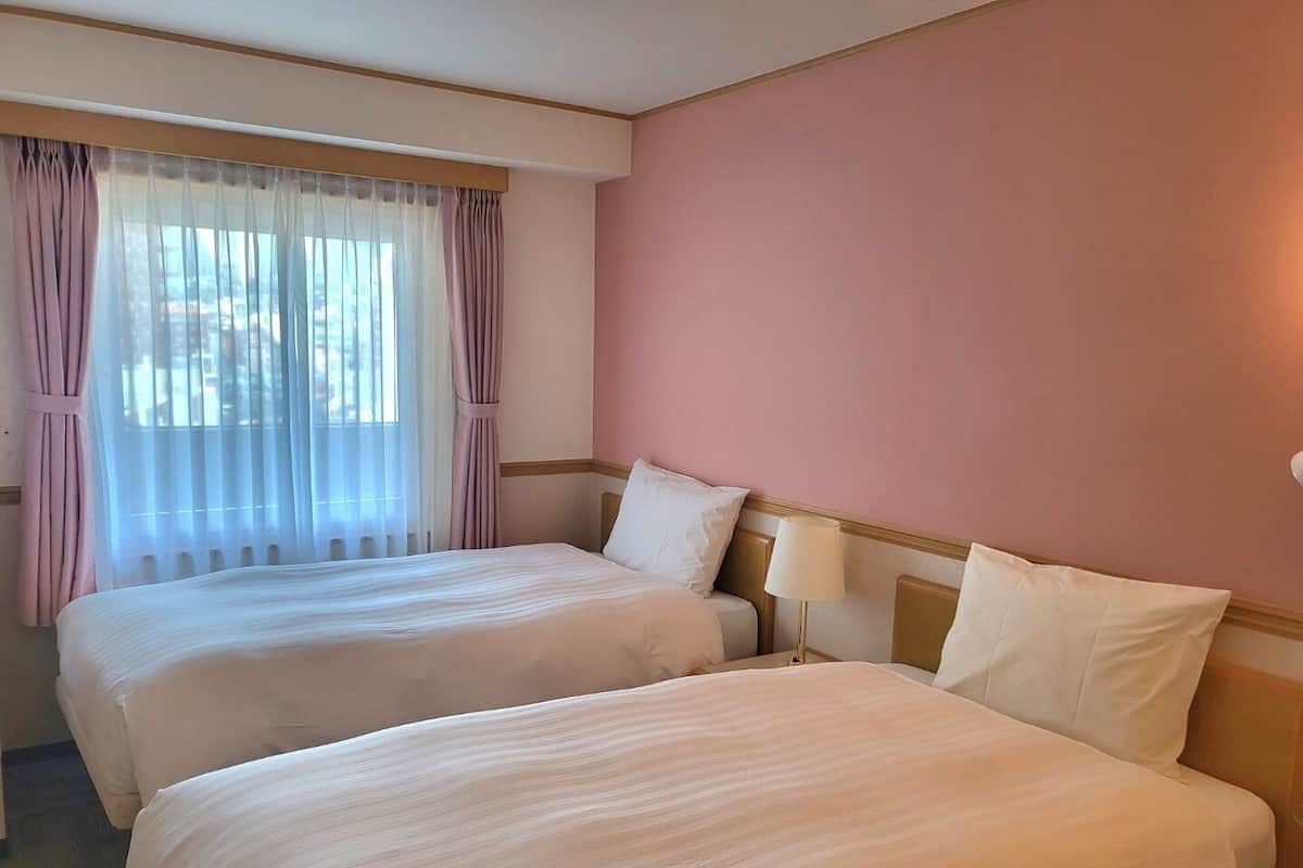 A hotel room with two beds, a large window with sheer curtains, and a pink wall.