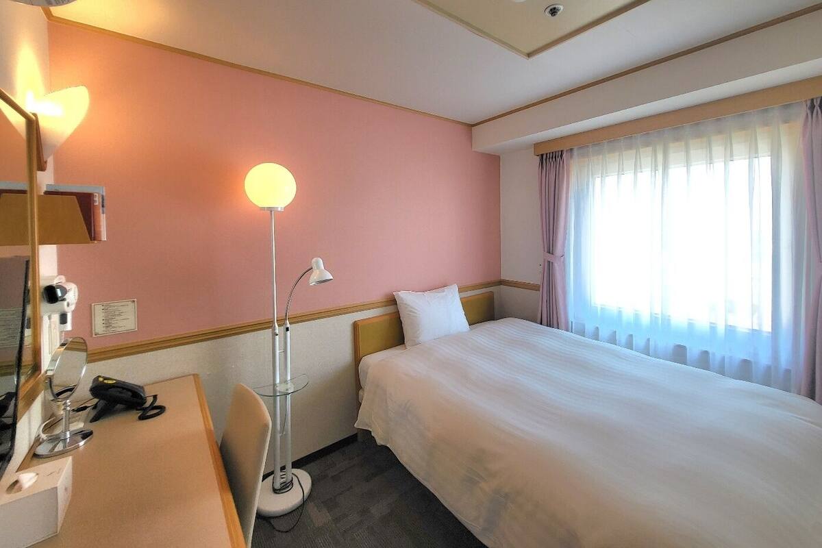 A hotel room with a bed, a desk with a lamp, a mirror, and a window with curtains.