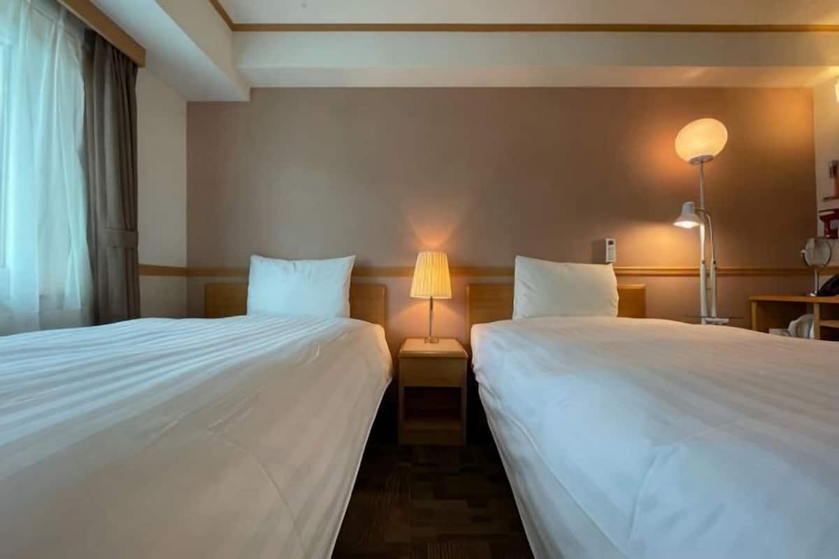 A hotel room with two beds, a nightstand, a lamp, and a wall-mounted lamp.