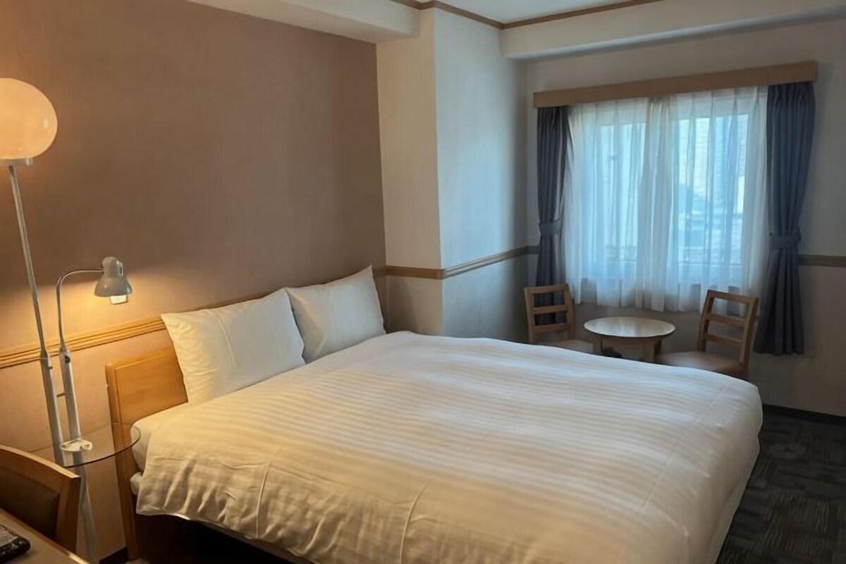 Ein Hotelzimmer mit einem großen Bett, einem Nachttisch mit Lampe, einem kleinen runden Tisch und einem Fenster mit Vorhängen.