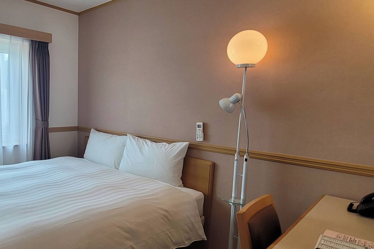 A hotel room with a bed, a desk, a lamp, and a window with curtains.