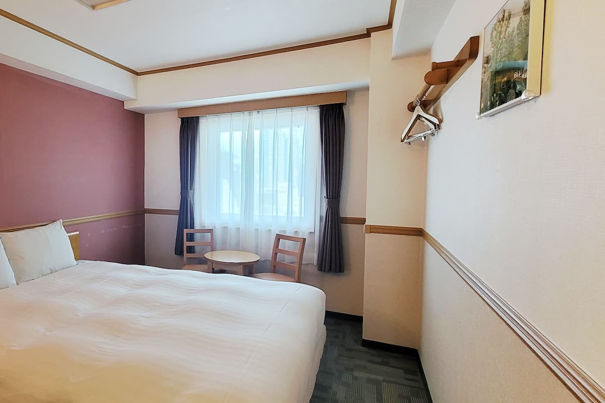 A hotel room with a large bed, a bedside table, a lamp, a window with curtains, and a small table with two chairs.