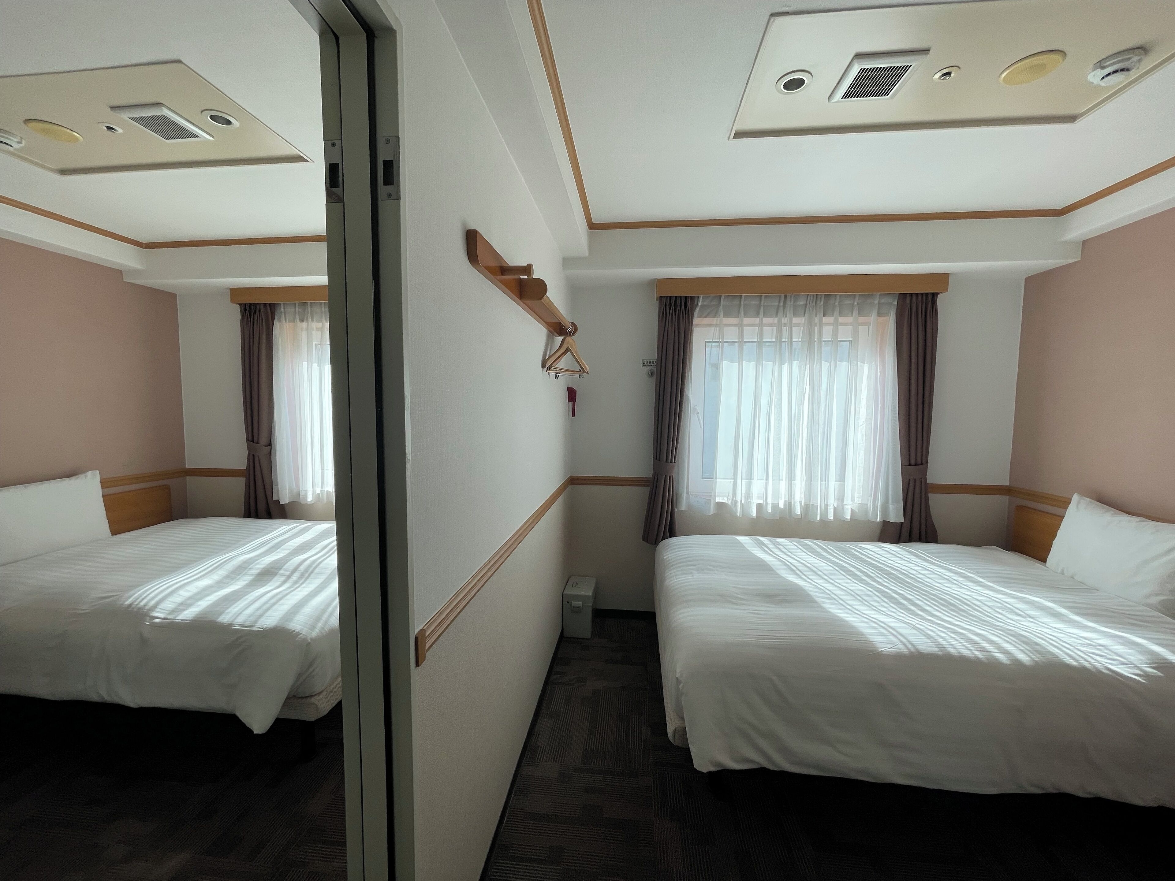 A hotel room with two beds, a mirror, a ceiling with ventilation, and a window with curtains.
