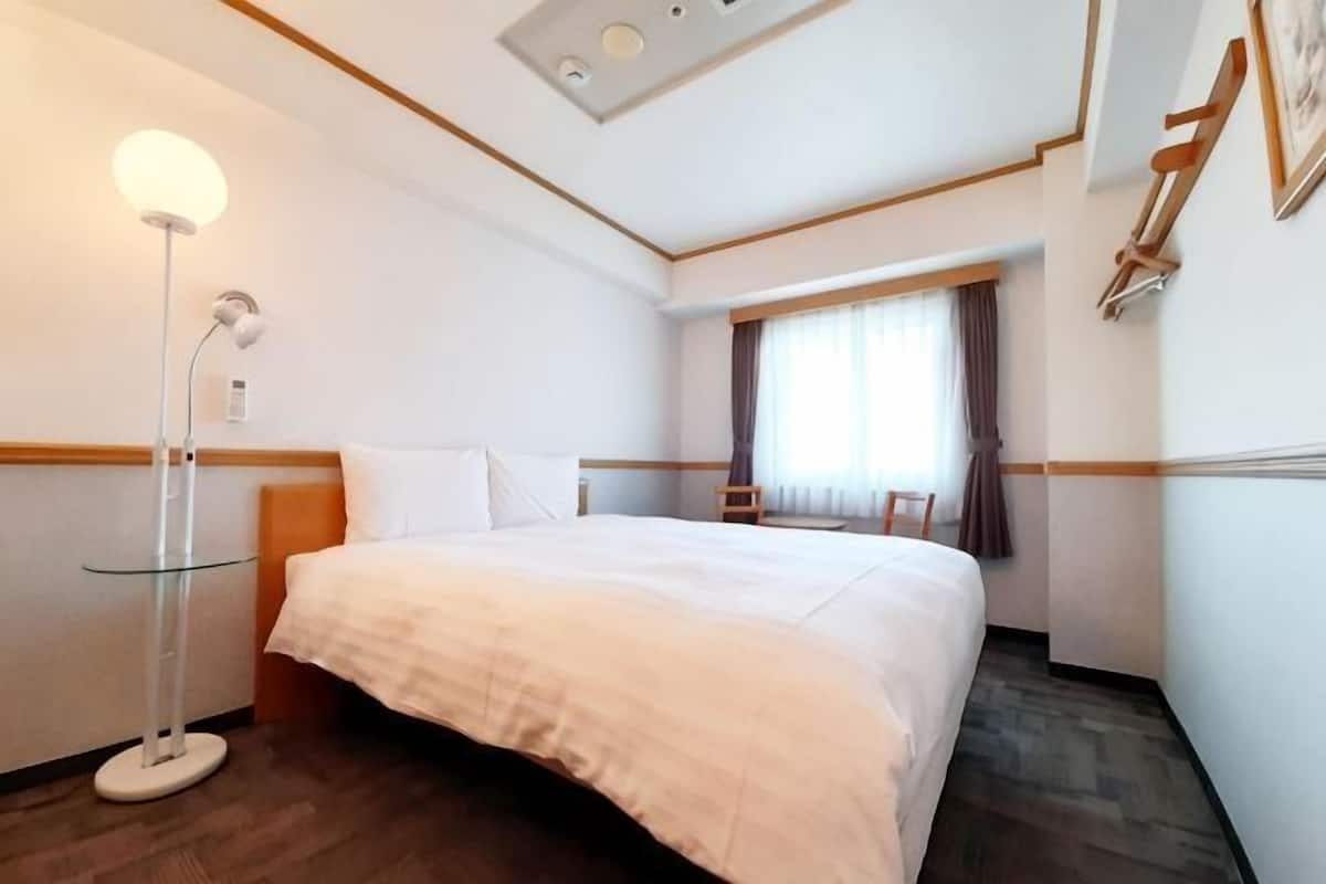 A hotel room with a bed, a bedside table, a lamp, a window with curtains, and a wooden headboard.