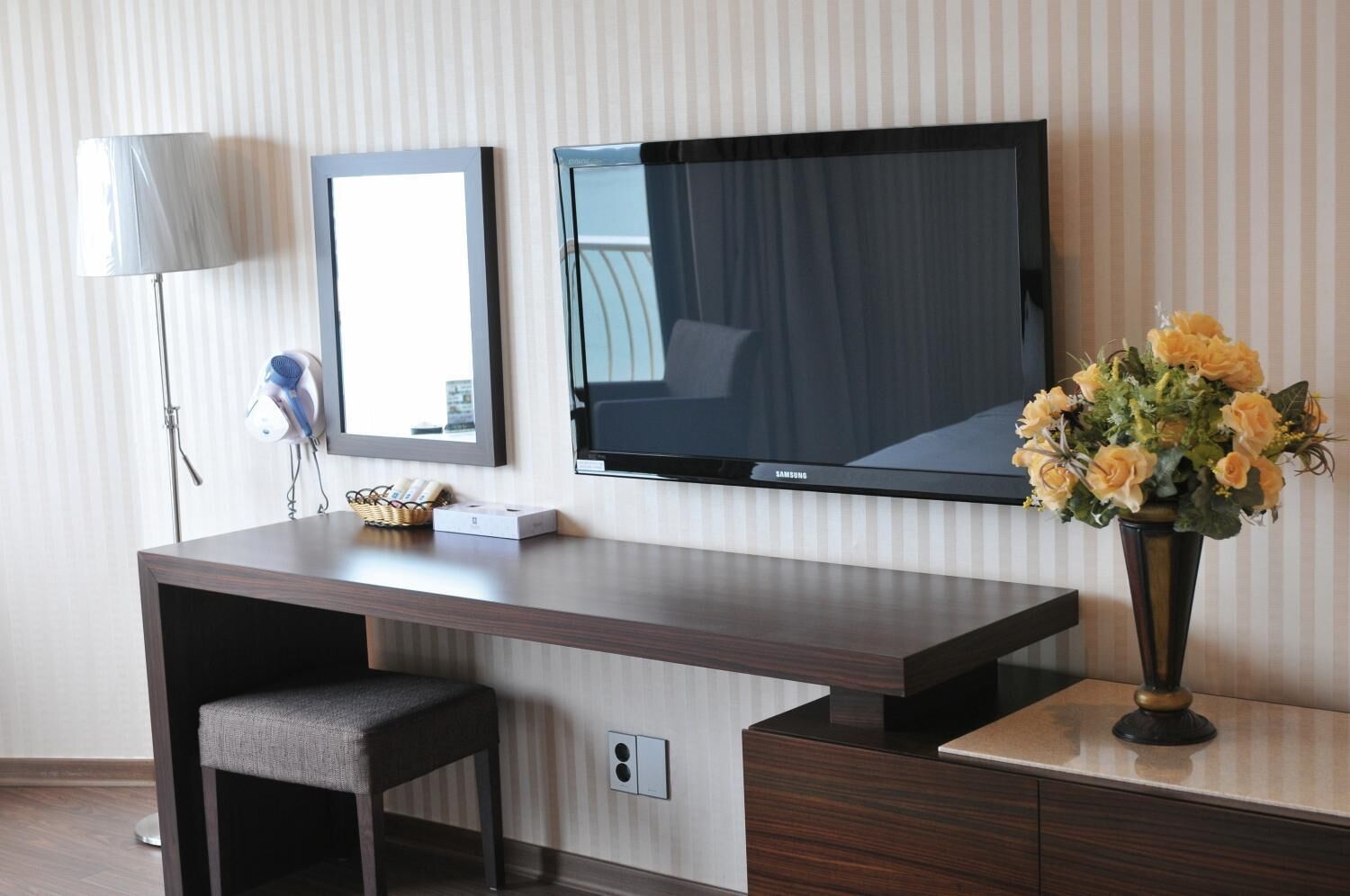 A modern hotel room with a flat-screen TV, a mirror, a desk with a chair, and a vase of flowers.