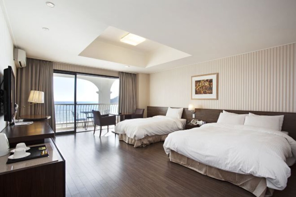 Ein Hotelzimmer mit zwei Betten, einem Schreibtisch, einem Stuhl, einem Balkon mit Blick und einem Bild an der Wand.