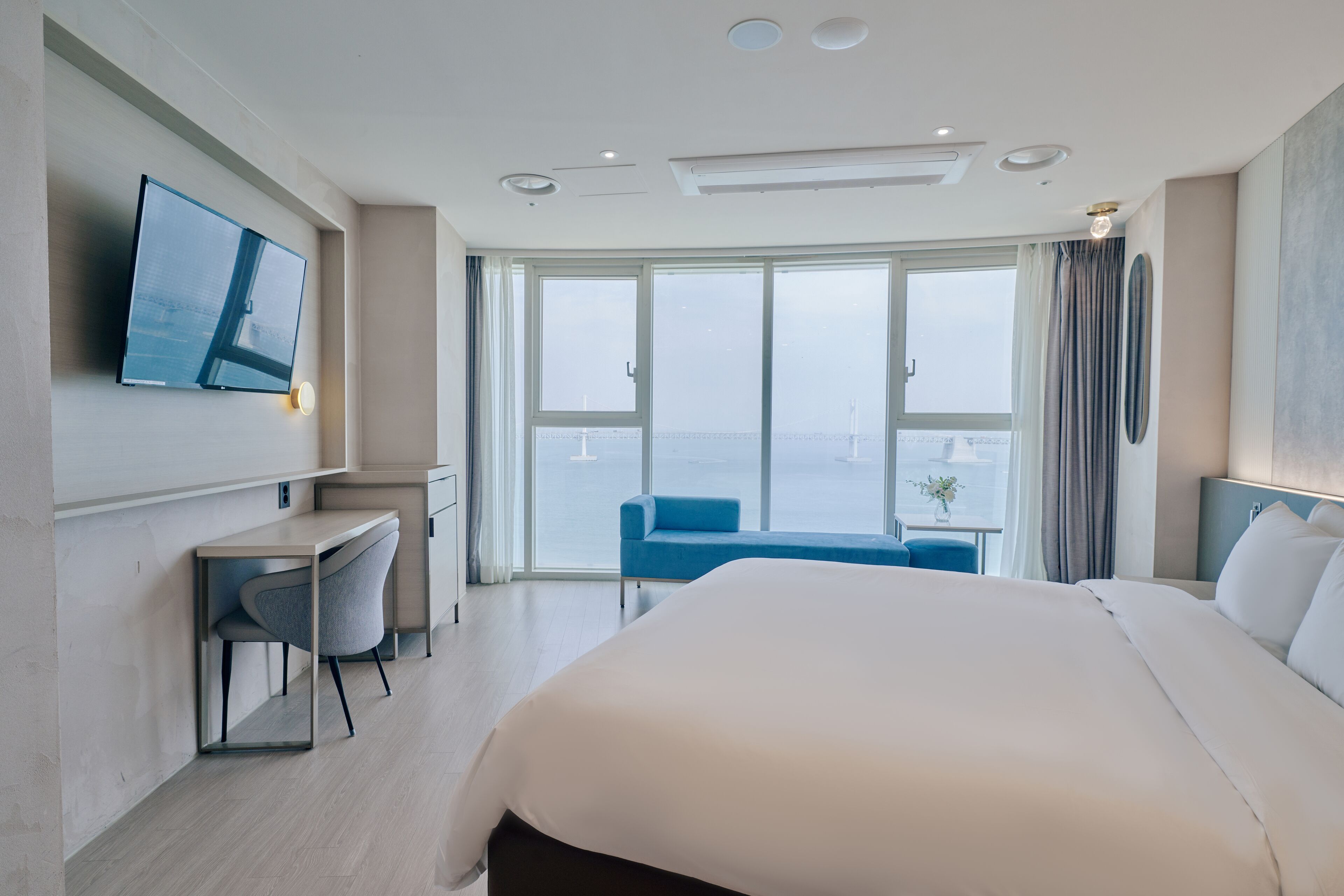 A hotel room with a large bed, a desk with a chair, a TV mounted on the wall, and a balcony with a view of the city.