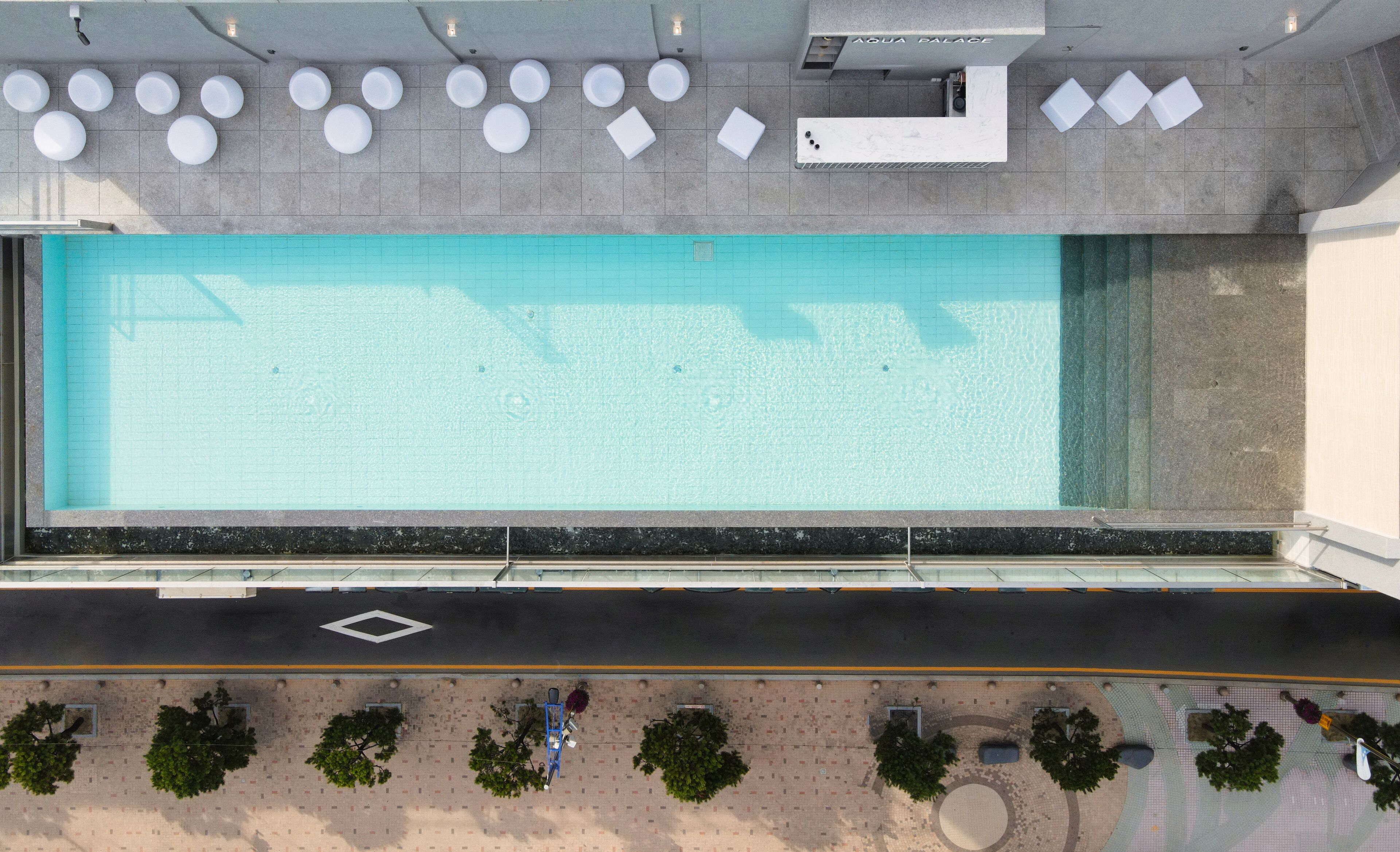 A rooftop swimming pool with a clear view of the sky, a lounge area with seating, and a road with trees and a pedestrian walkway.
