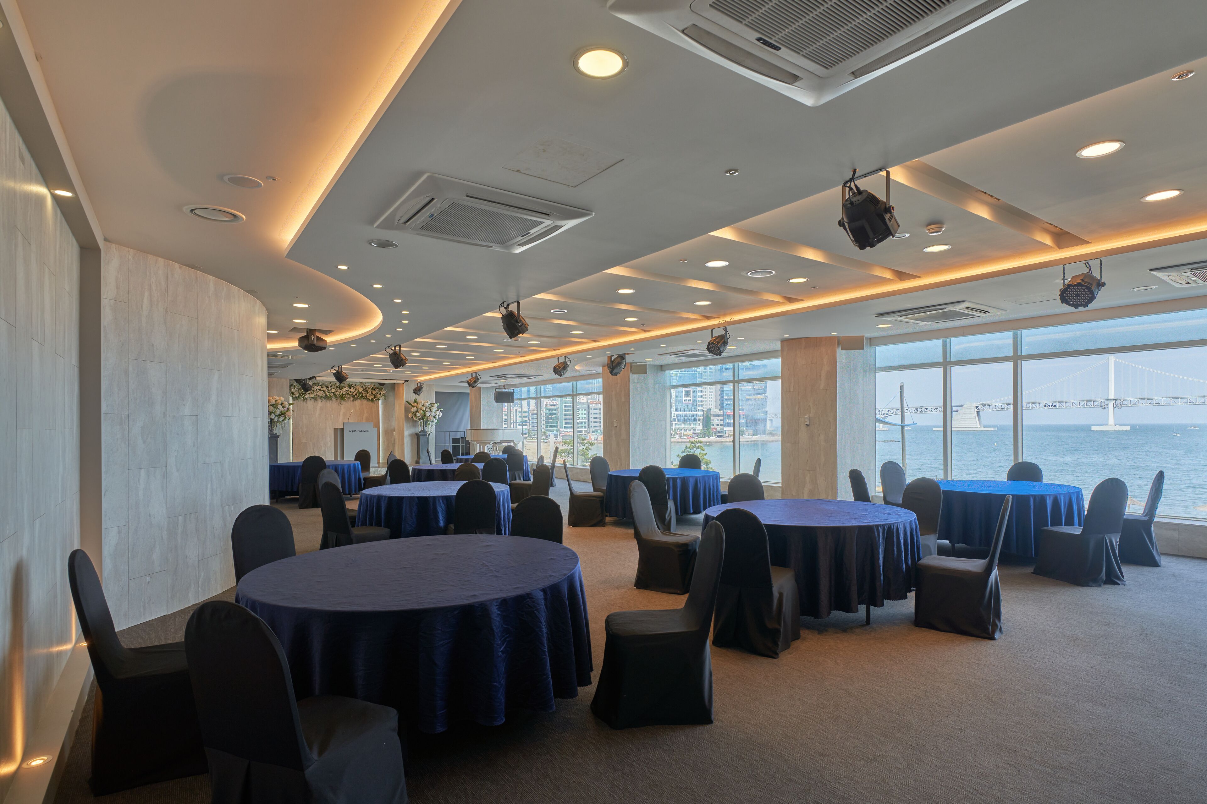 A conference room with round tables and chairs, large windows with a view of a bridge and water, and a modern ceiling with recessed lighting.