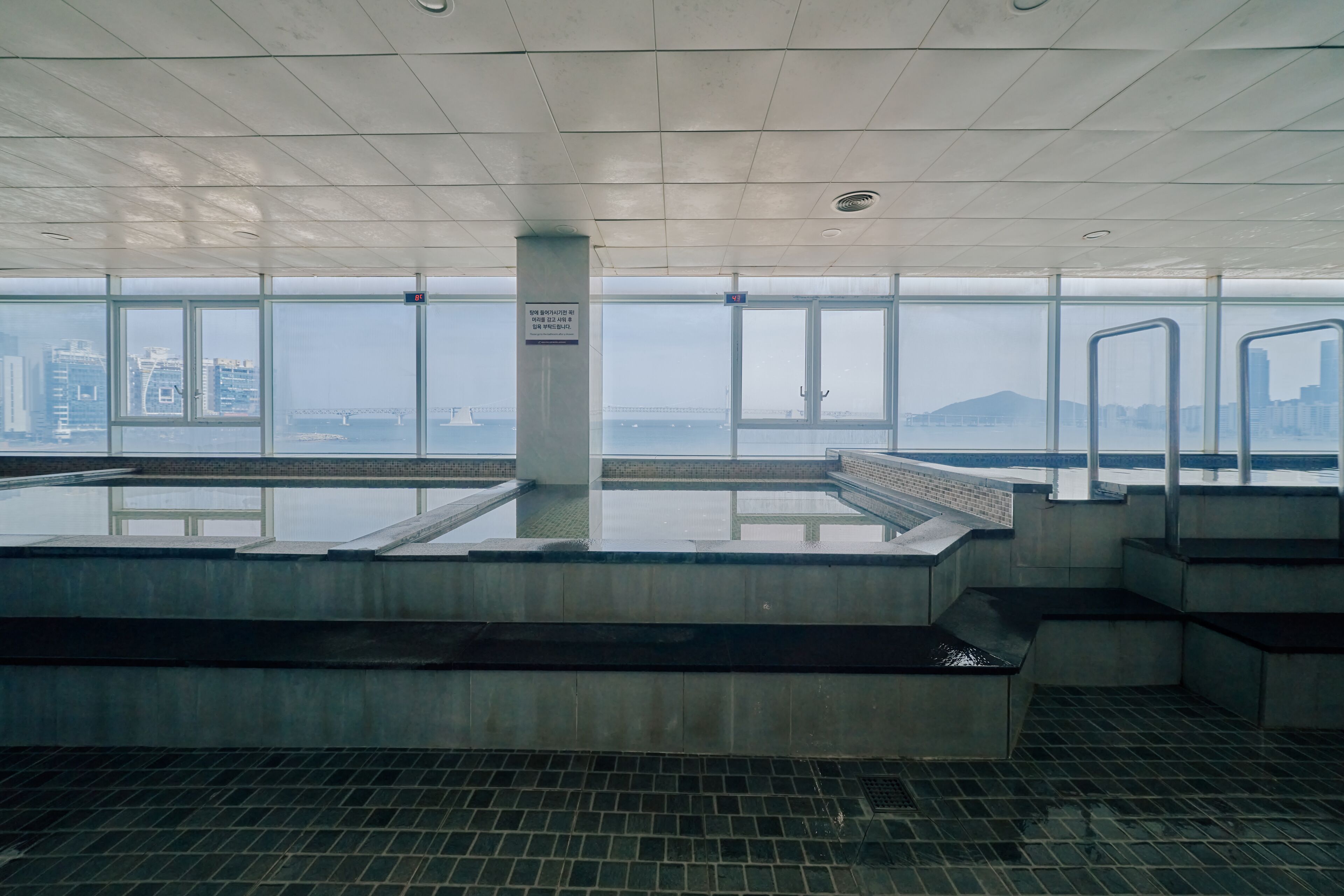 A modern indoor swimming pool with clear glass walls, a tiled floor, and a view of a cityscape and bridge.