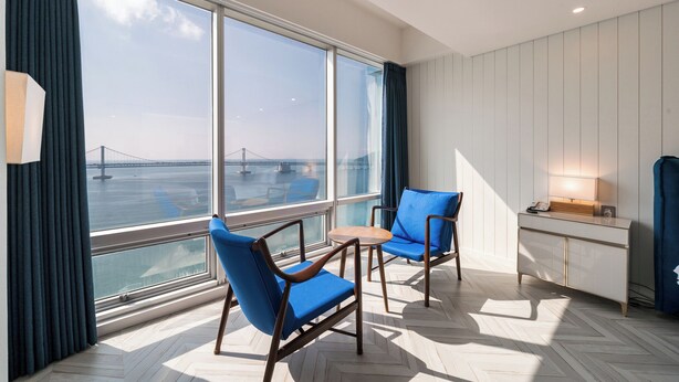 Ein Zimmer mit großen Fenstern, Blick auf eine Brücke, zwei blaue Stühle, ein kleiner Tisch und eine weiße Kommode.