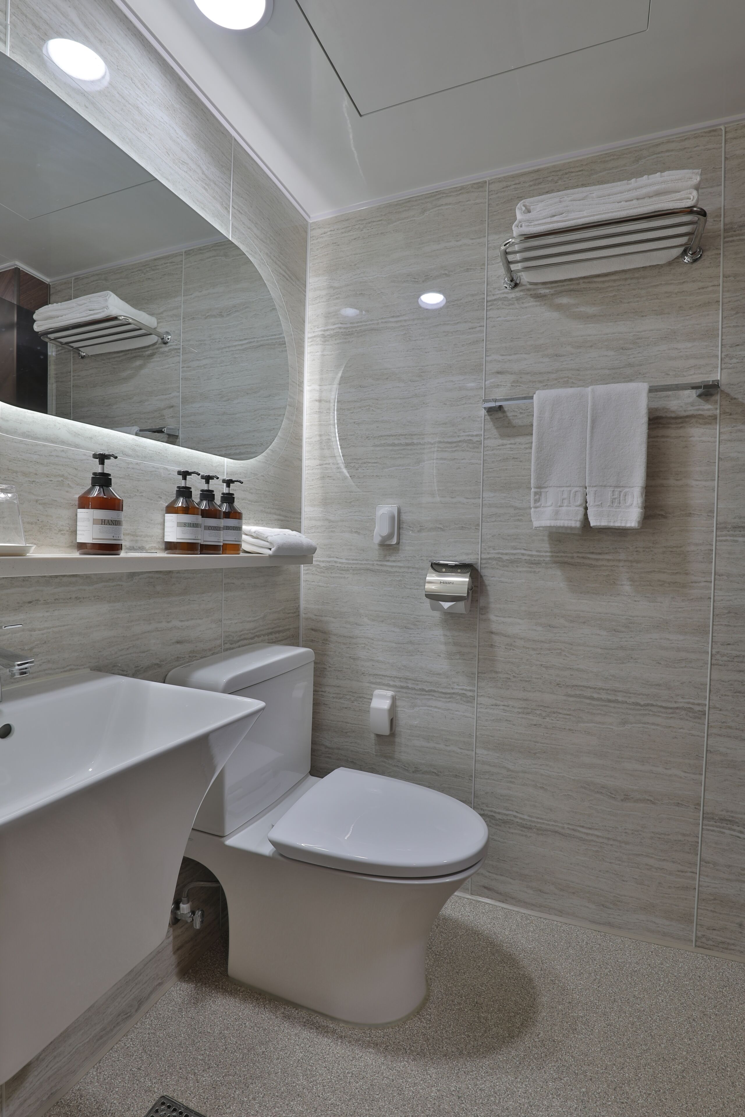 Une salle de bain moderne avec un lavabo, des toilettes et une douche, le tout de couleur blanche. On peut voir des flacons de produits de toilette sur le comptoir, et des serviettes sont suspendues à un porte-serviettes.