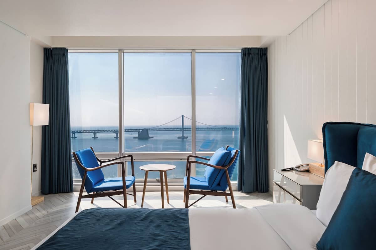 A hotel room with a large window, two blue chairs, a bed, a bedside table with a lamp, and a view of a bridge.