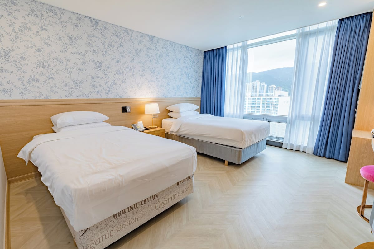 A hotel room with two beds, a wooden headboard, a nightstand with a lamp, and a view of buildings through large windows.