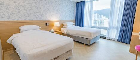 Quarto de hotel com duas camas, cabeceira de madeira, mesa de cabeceira com abajur e vista para edifícios através de grandes janelas.