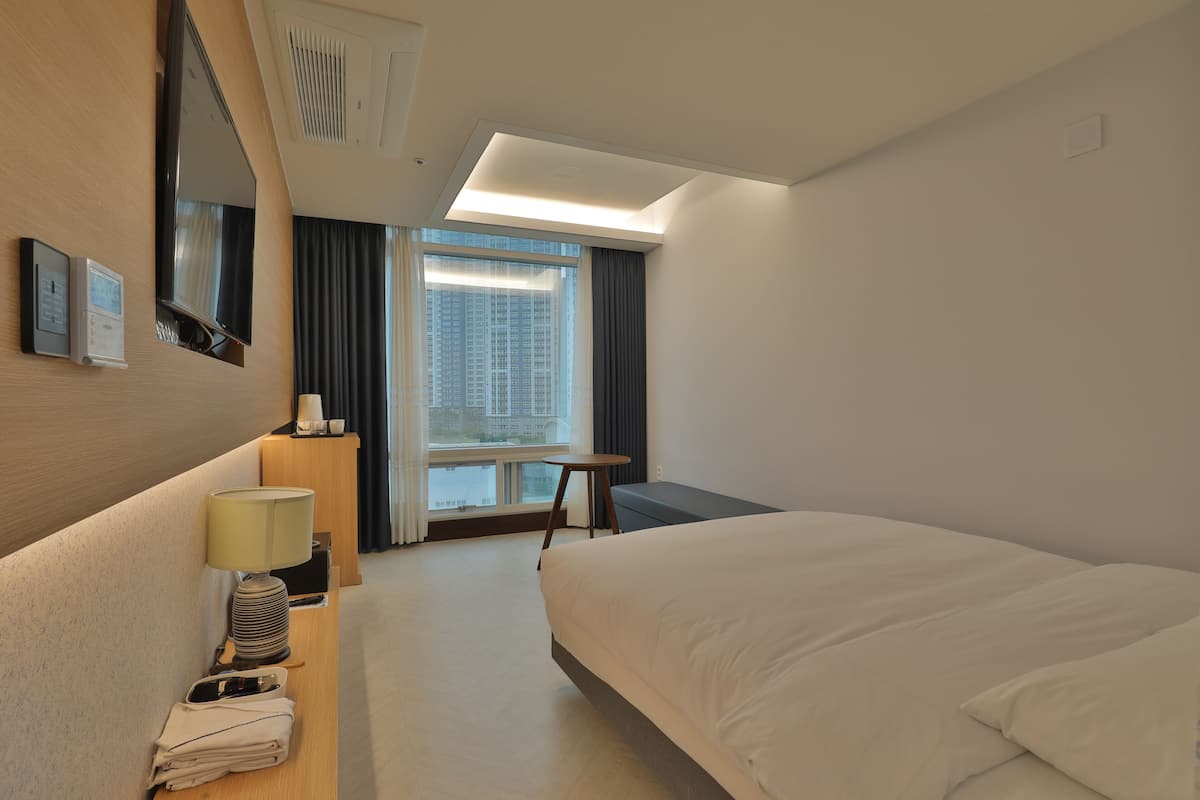 A hotel room with a bed, a desk with a lamp, a small table, a window with curtains, and a view of buildings outside.