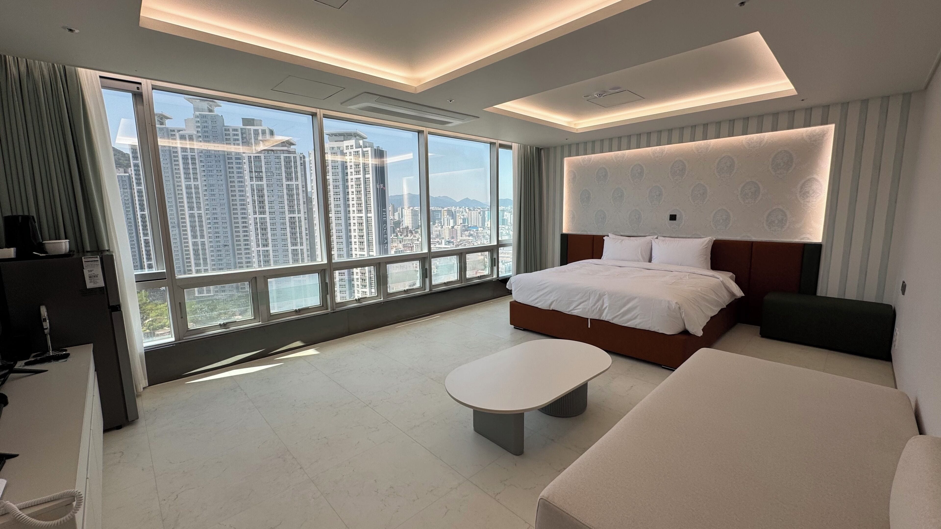 A modern hotel room with a large bed, a small table, and a sofa. There is a large window offering a city view.